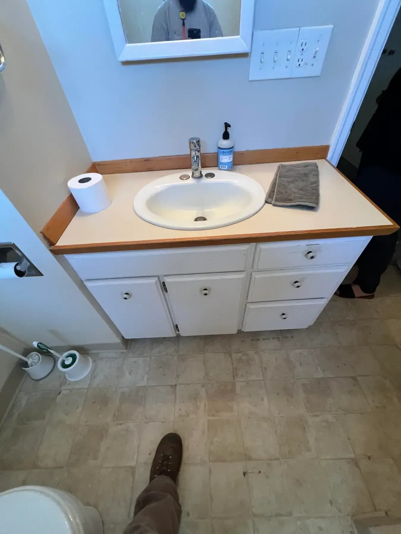 Small bathroom vanity with sink, mirror, toiletries, toilet paper, and tiled floor; a shoe is visible at the bottom.