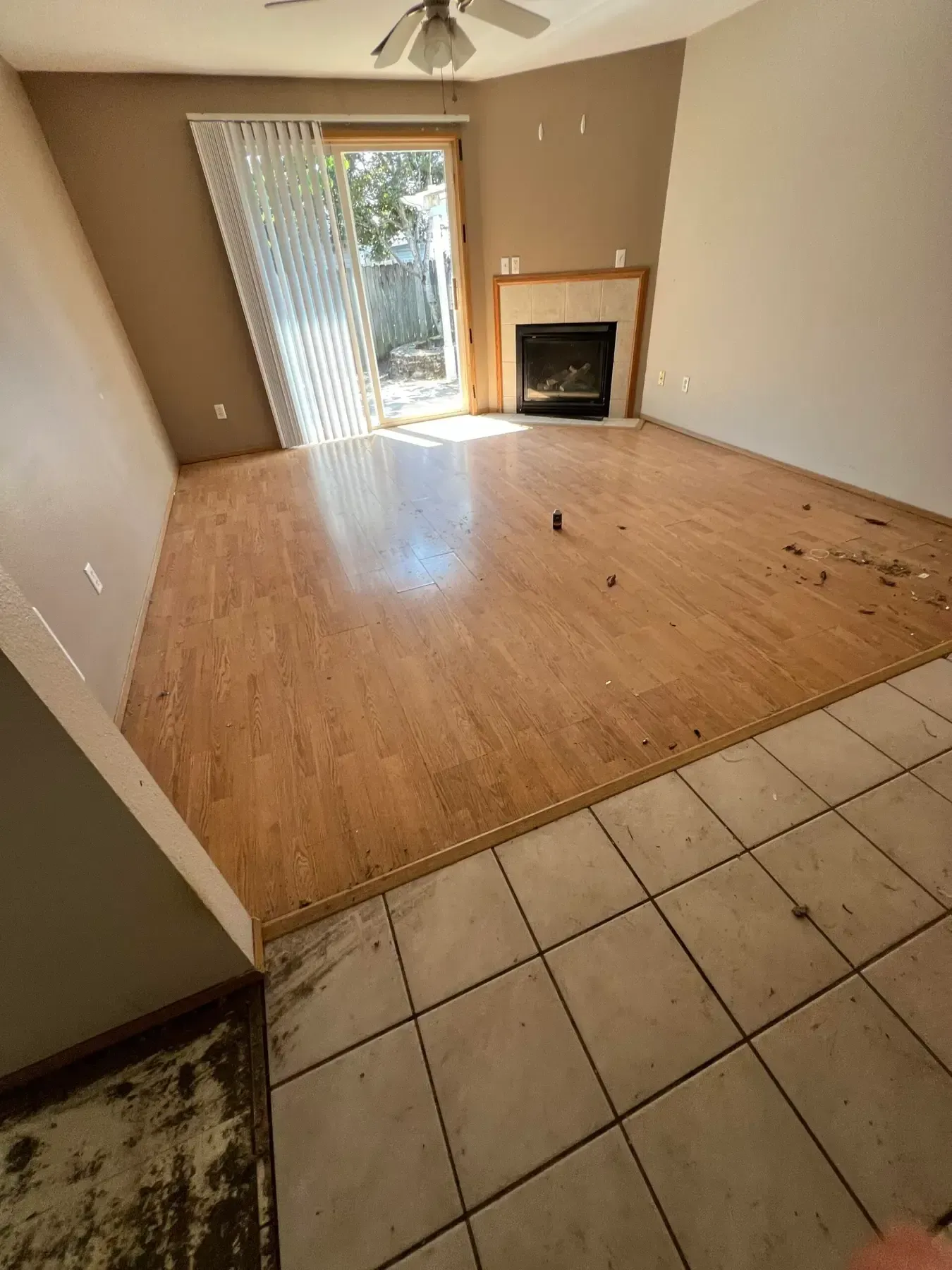 Empty living room with tan walls, tile and wood floors, a fireplace, and sliding glass door with vertical blinds