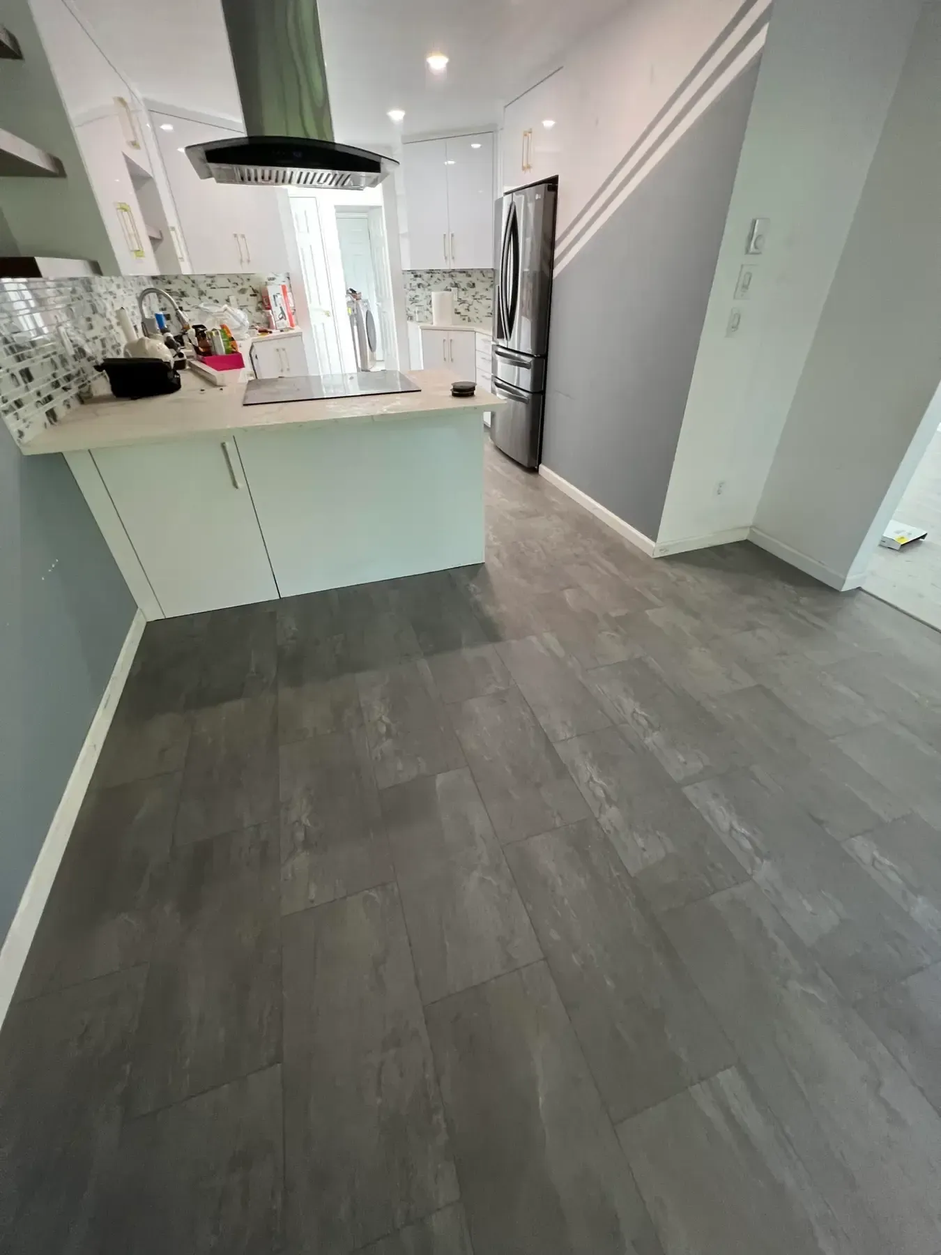 Modern kitchen with white island, gray wood floor, stainless fridge, and staircase in the background