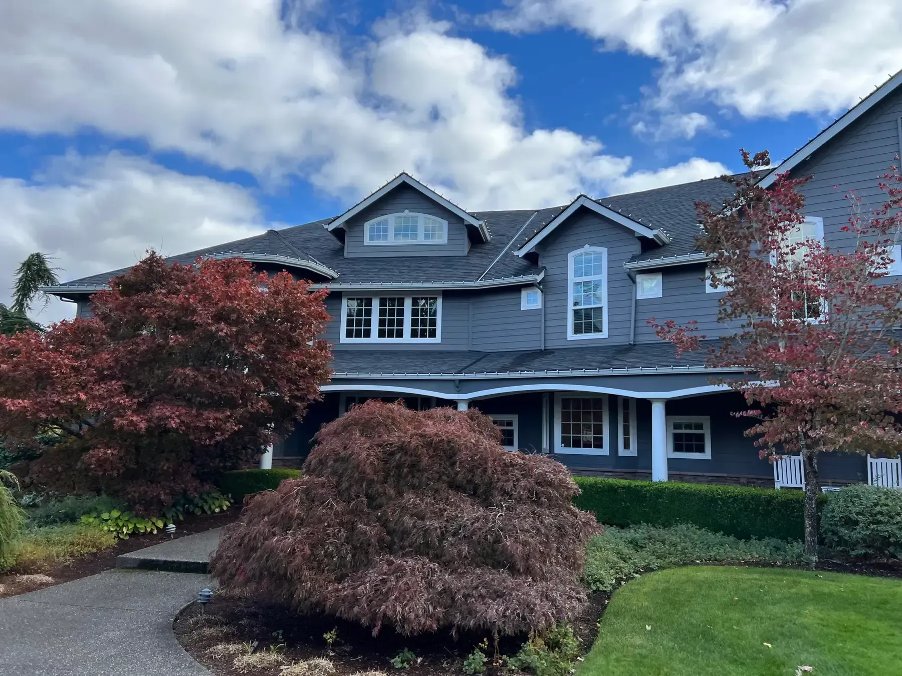 Large gray house with gabled roof, red shrubs, and cloudy blue sky above a front lawn