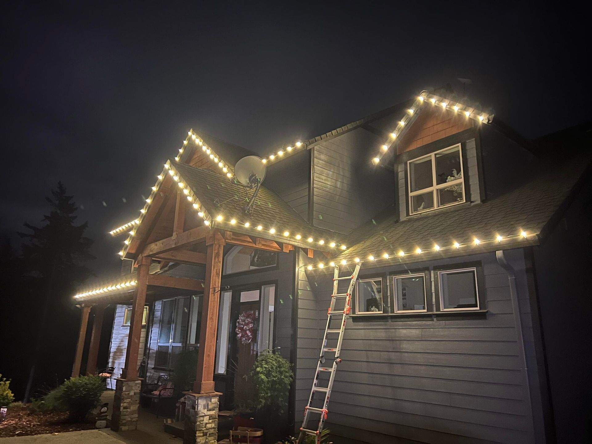 House outlined with warm white string lights at night, with a ladder leaning against the roof.