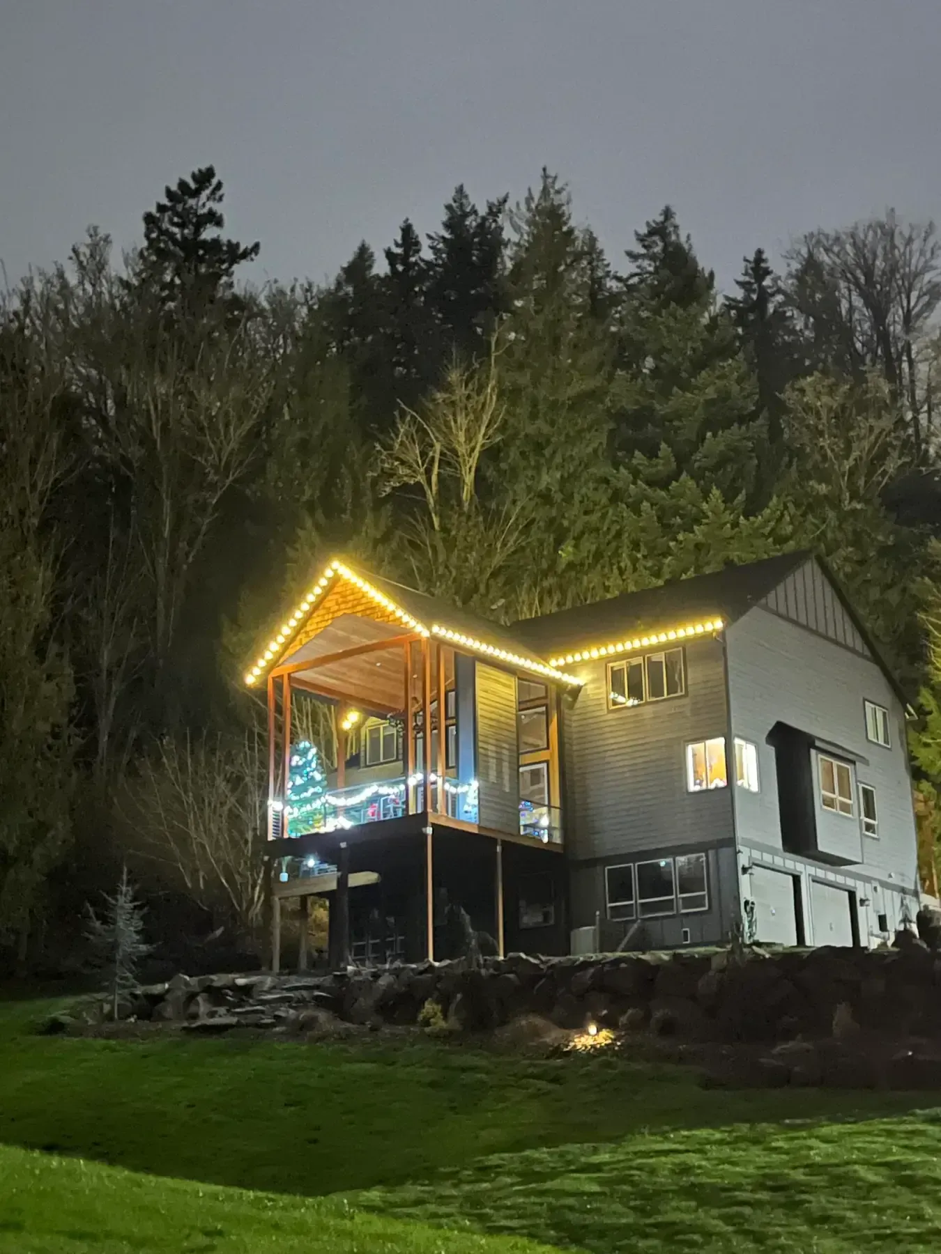 Lit house at night on a wooded hillside with glowing roofline and porch lights