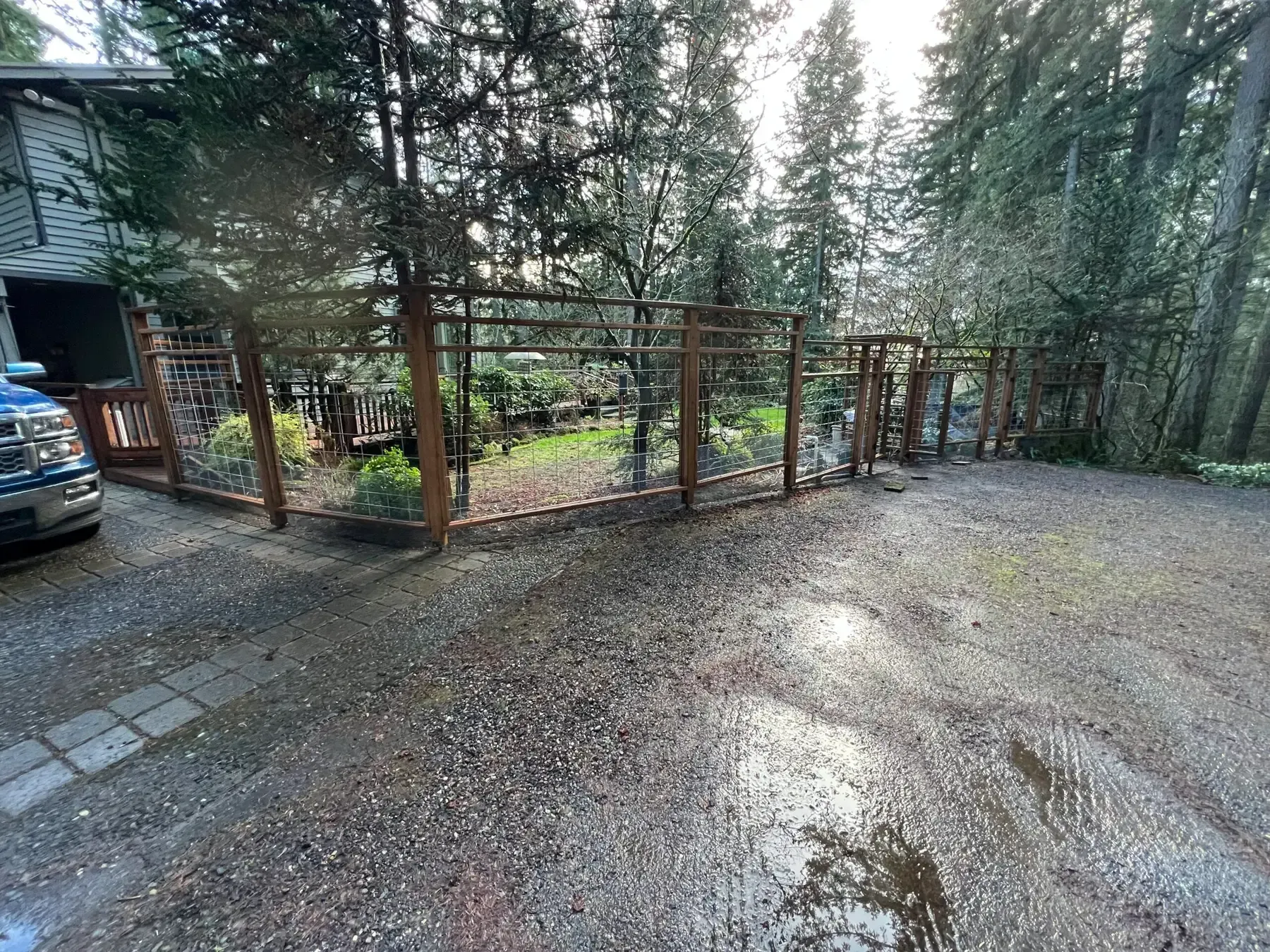 Gravel driveway beside a house, fenced garden, and tall evergreen trees in a wooded setting.