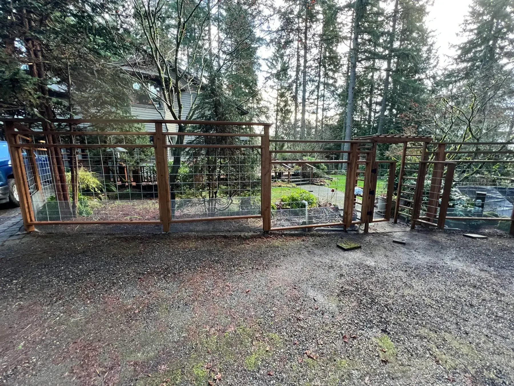 Rusty metal fence and gate beside a forested path, with gravel foreground and tall evergreen trees beyond.