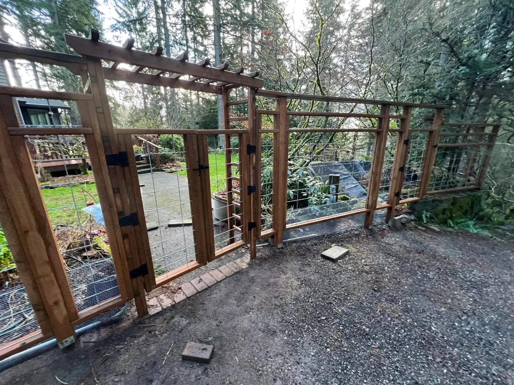 Wooden fence and gate beside a forested creek, with gravel ground and tall trees beyond.