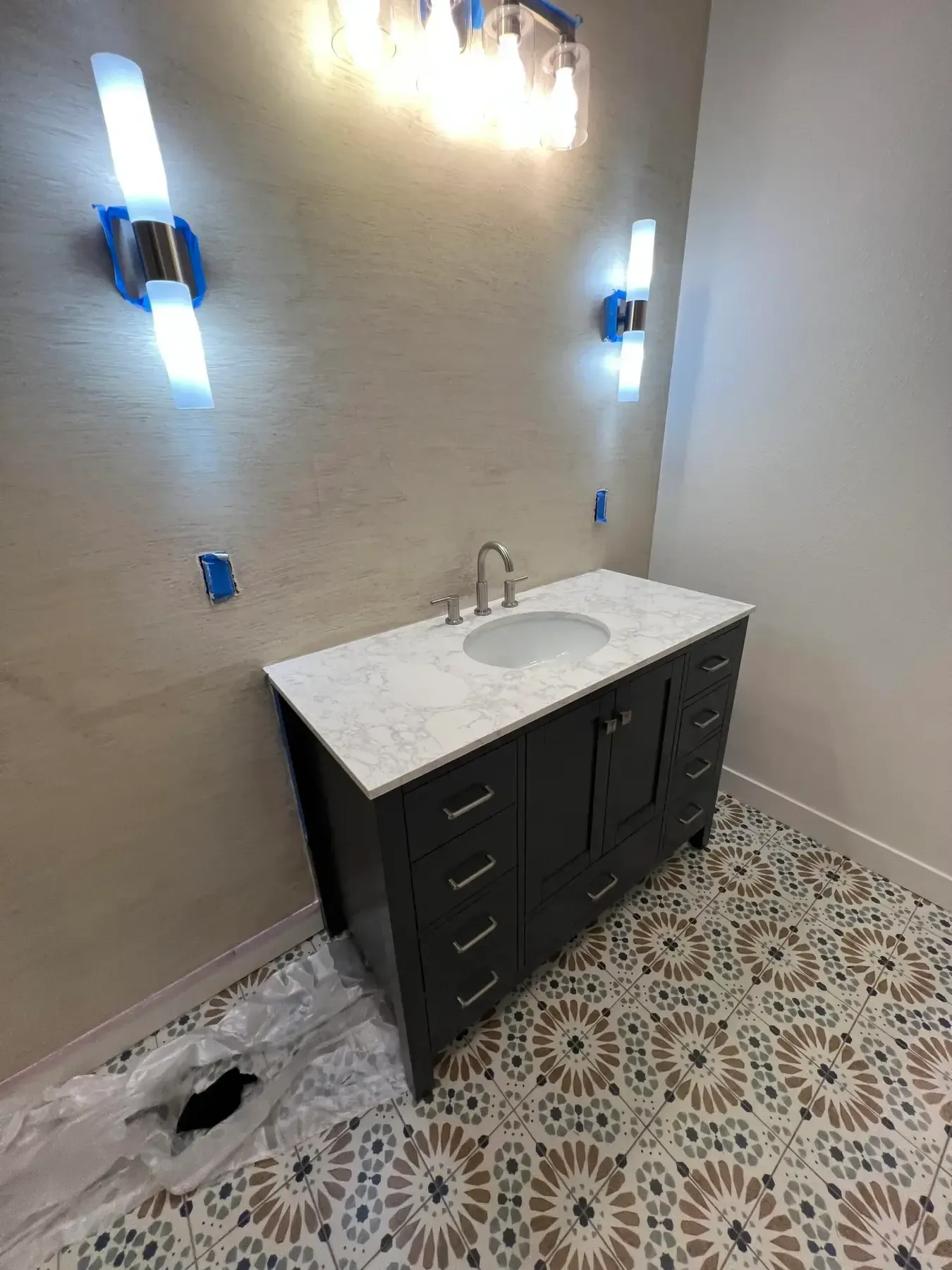 Bathroom vanity with white countertop, dark cabinets, wall sconces, and patterned tile floor under renovation