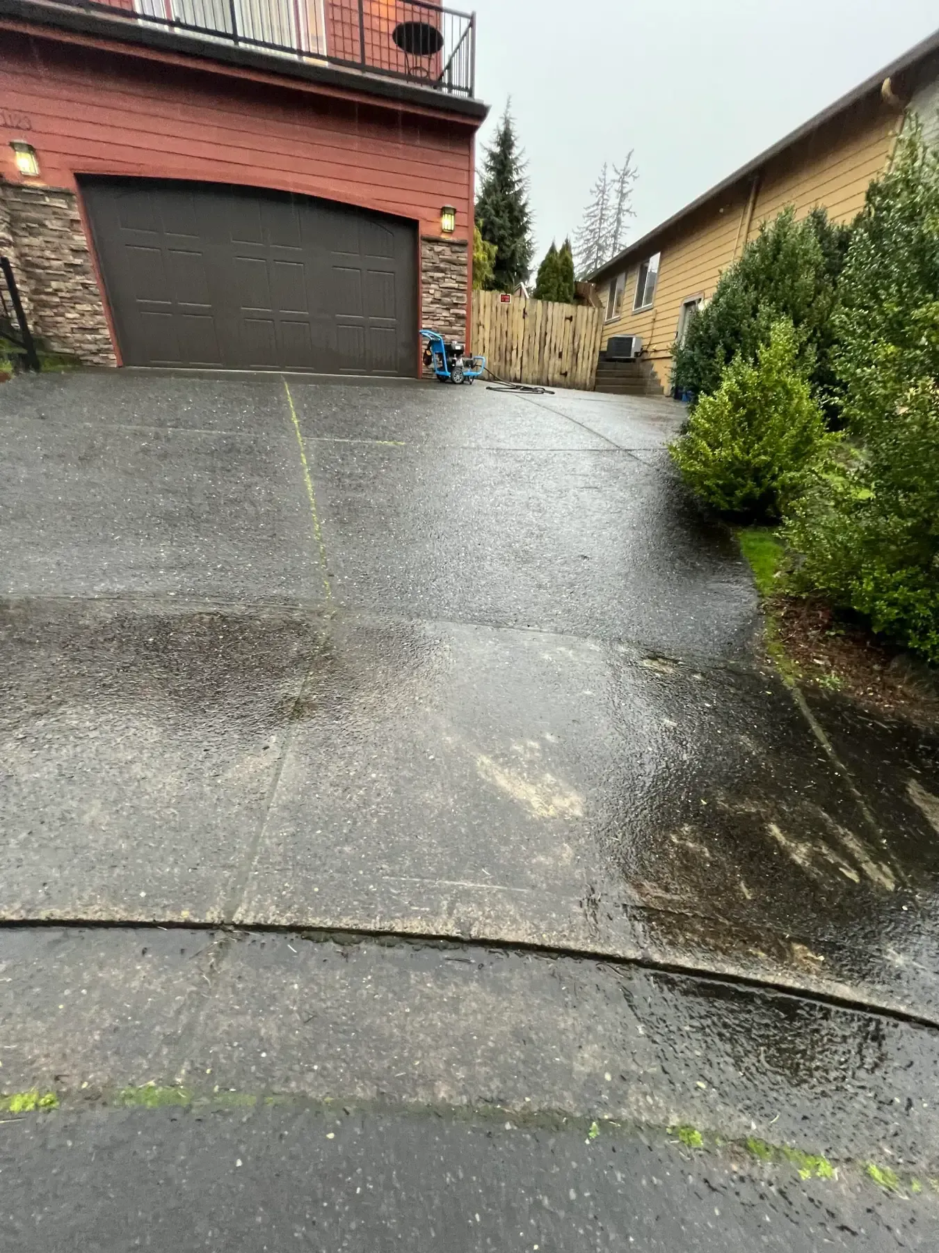 Wet sloped driveway beside a red garage and hedges on a rainy day