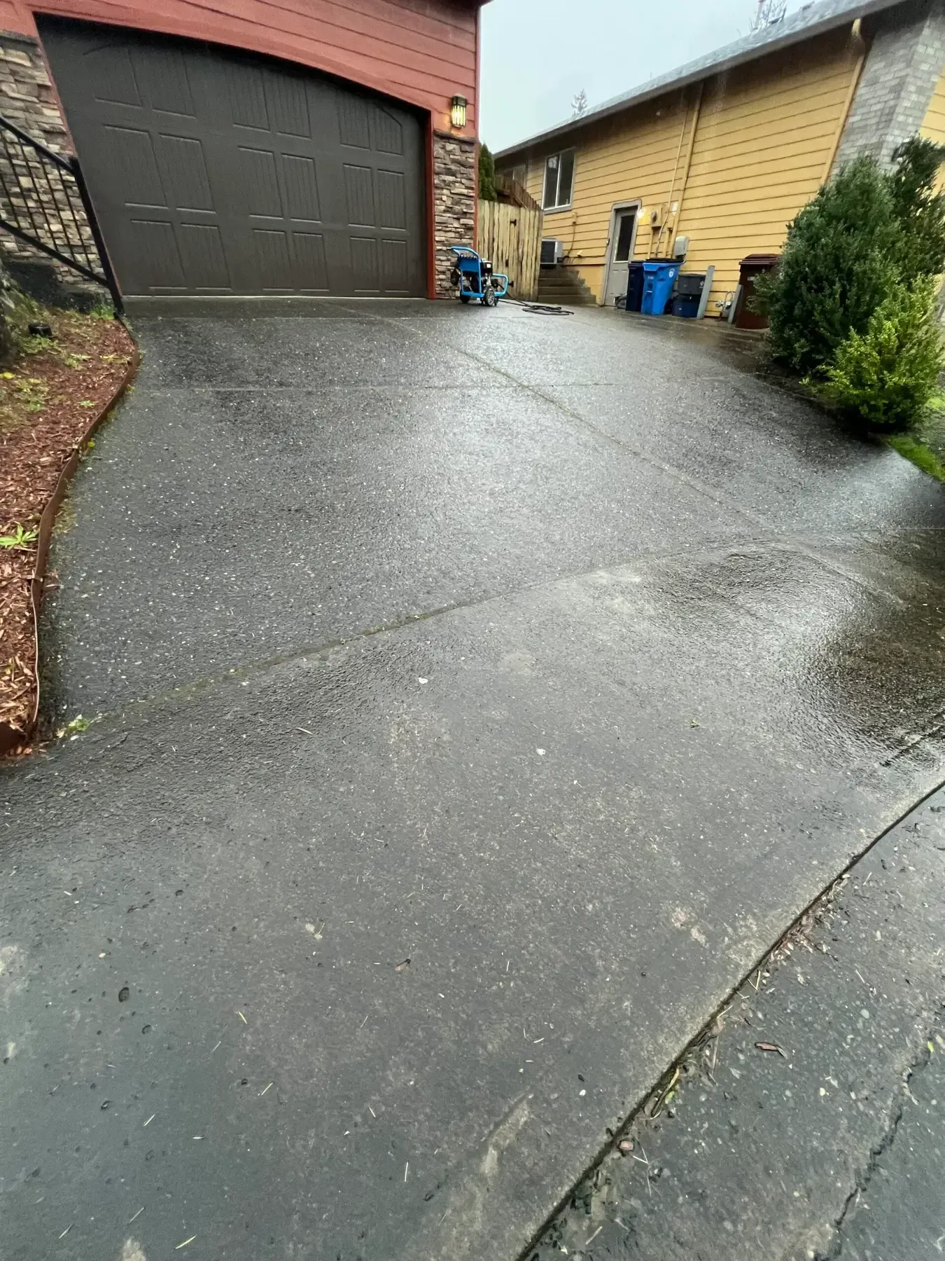 Wet driveway leading to a garage beside a house, with recycling bins and shrubs on the right.