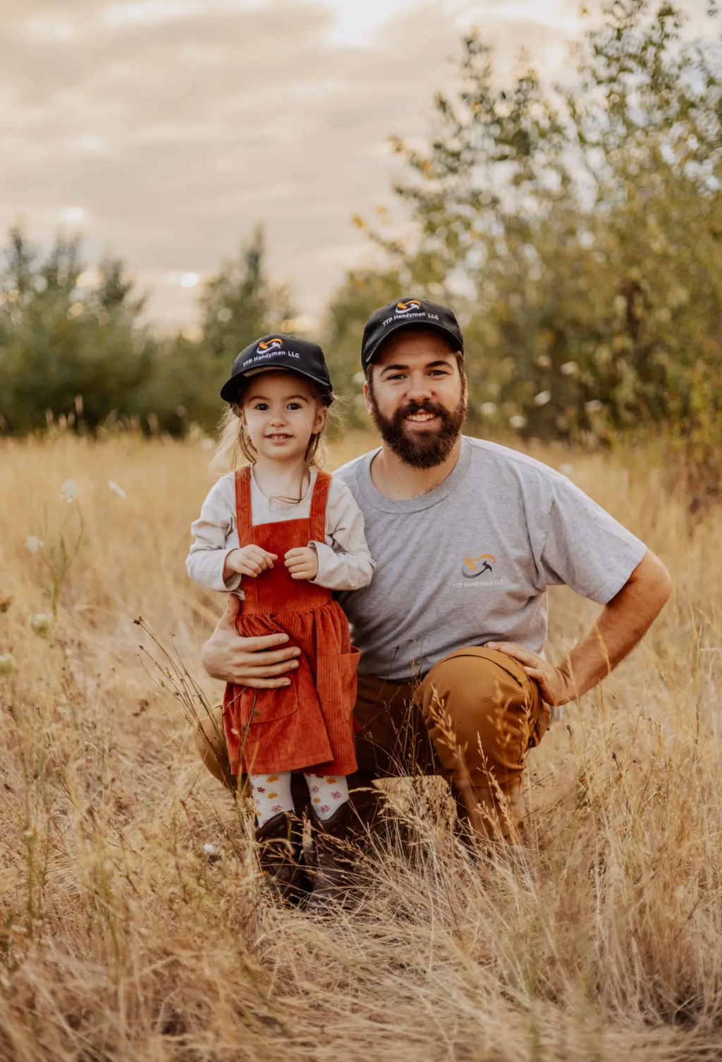 Child in red overalls and adult kneeling in a grassy field, both wearing caps and smiling.