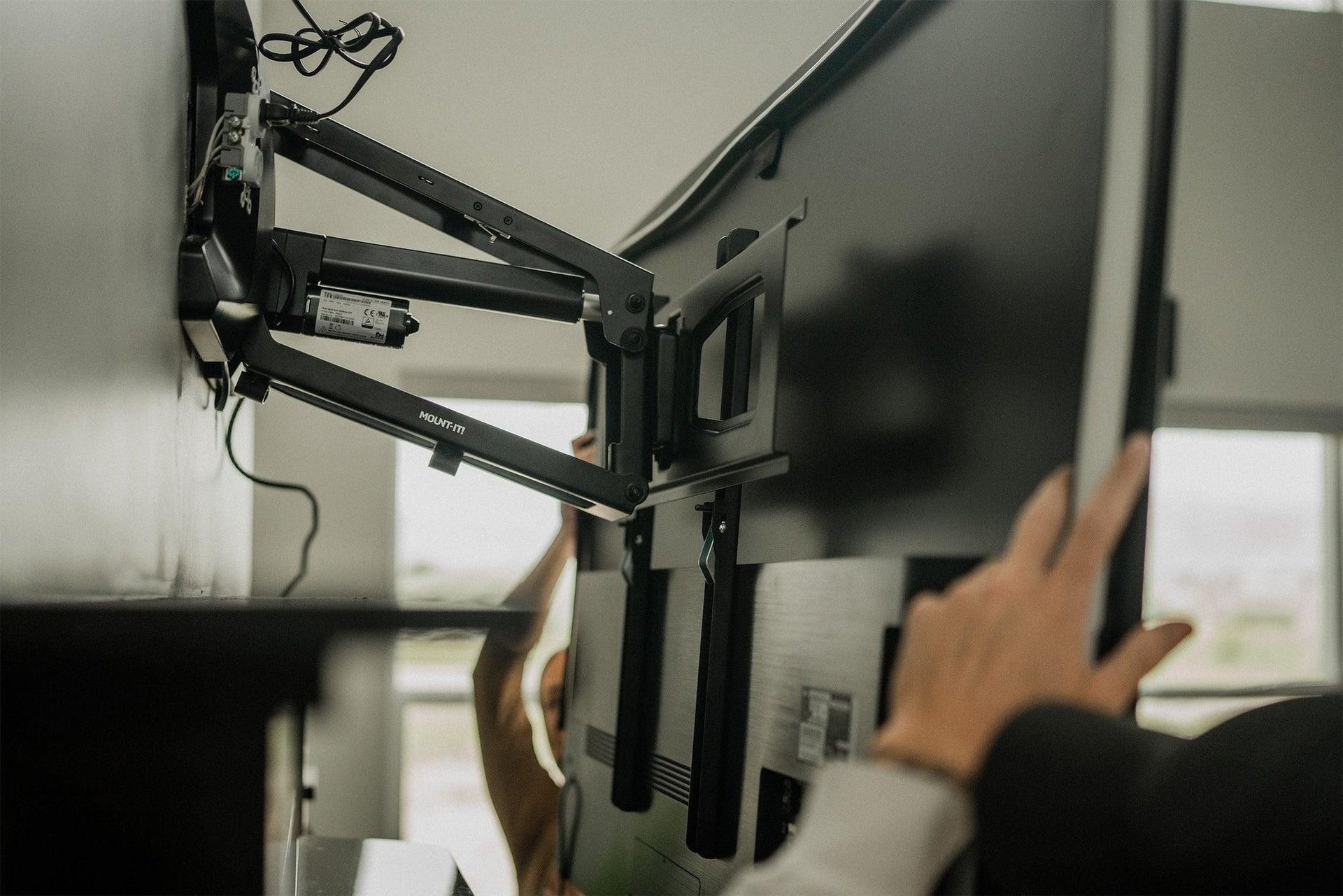 A person adjusting a black monitor arm in a dim office workspace