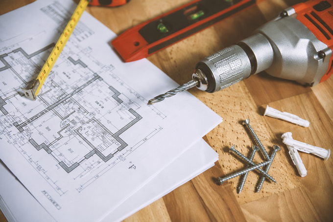 Drill, tape measure, and blueprint on a wooden table with screws and a level, suggesting a home renovation project