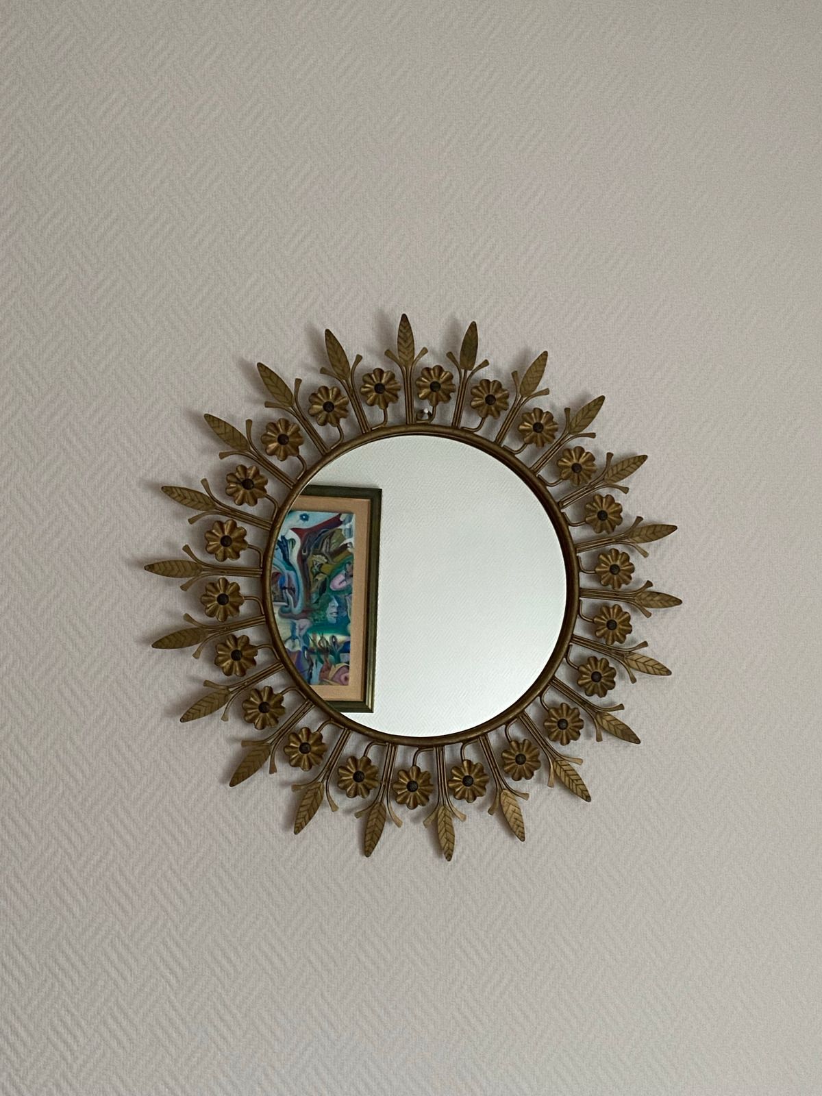 Round gold sunburst mirror on a beige wall, reflecting a framed artwork.