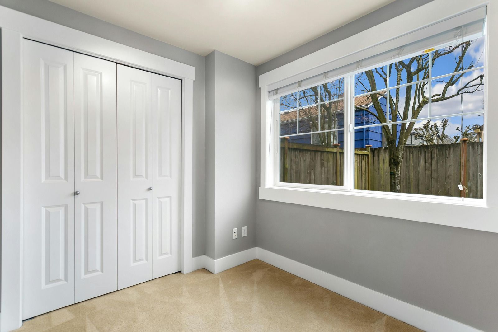 Empty bedroom corner with white closet doors, gray walls, beige carpet, and a window looking outside