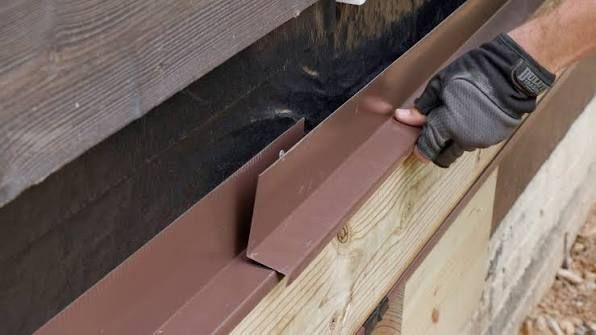 Gloved hand installing a brown metal trim strip along the edge of a wooden roof or wall.