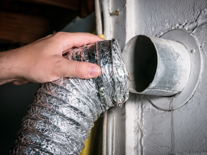 A hand attaching a flexible silver duct to a round metal vent pipe in a wall.