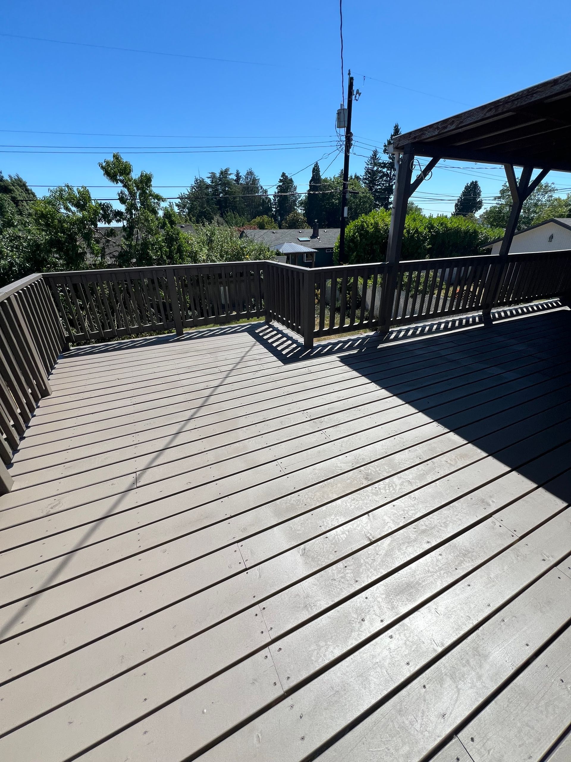 Sunny outdoor deck with gray wood planks, black railing, and a covered patio overlooking trees and sky