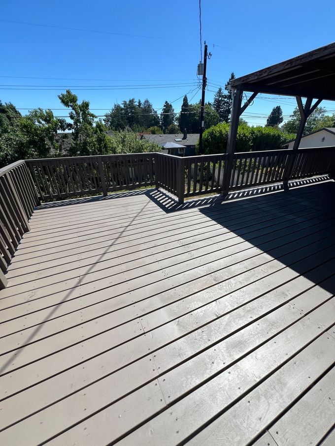 Sunny outdoor deck with gray composite boards, black railing, and a partial roof overlooking trees and sky