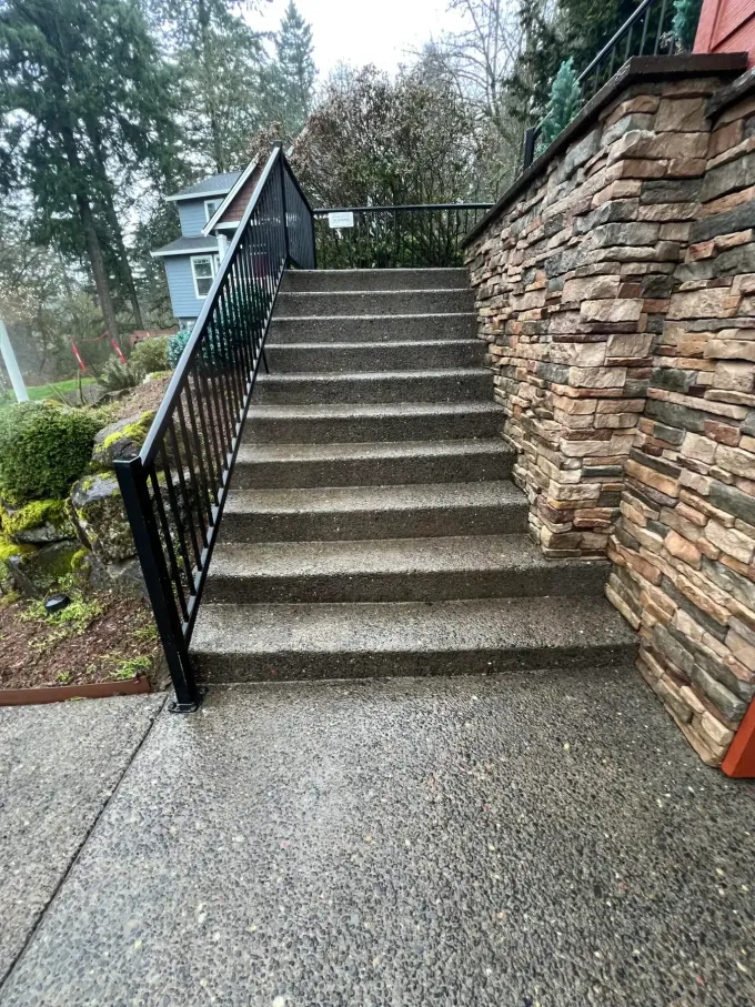 Concrete stairs beside a stone wall and black railing, leading up a hillside in a wooded area.