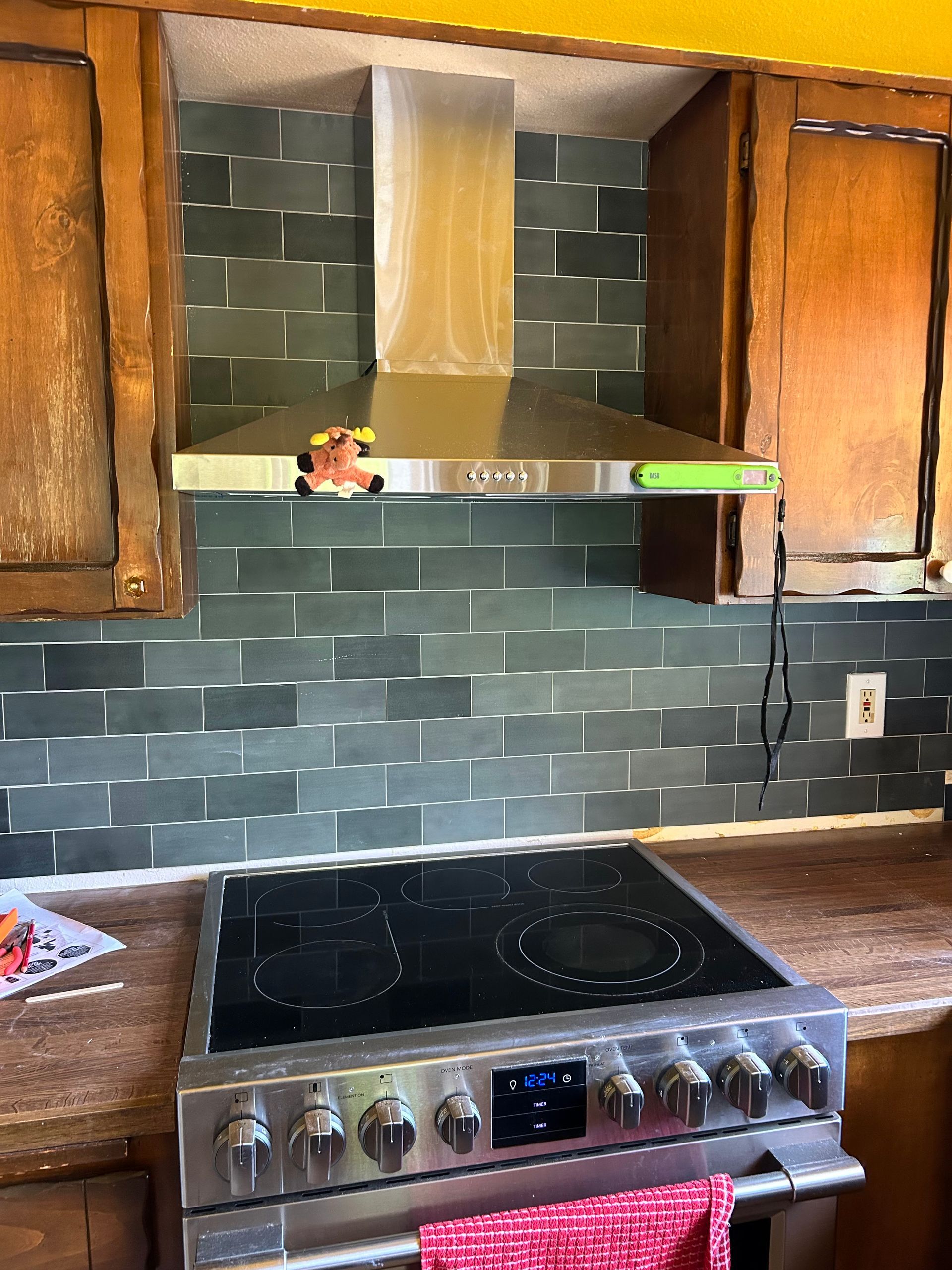 Kitchen stove beneath tiled backsplash and range hood, with wooden cabinets and a floating shelf above.