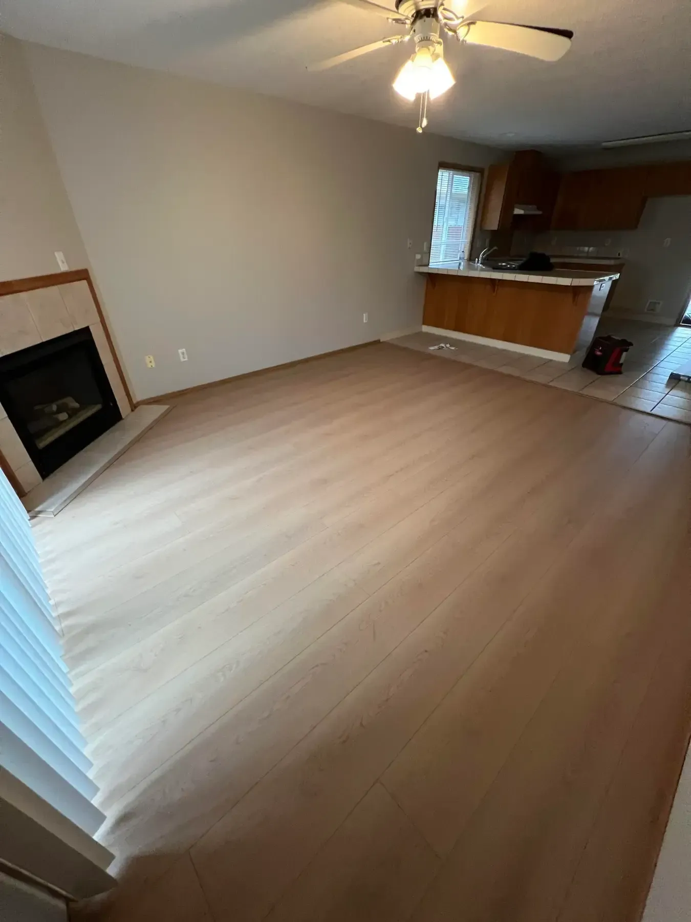 Empty living room with light wood floor, fireplace, ceiling fan, and kitchen counter in back