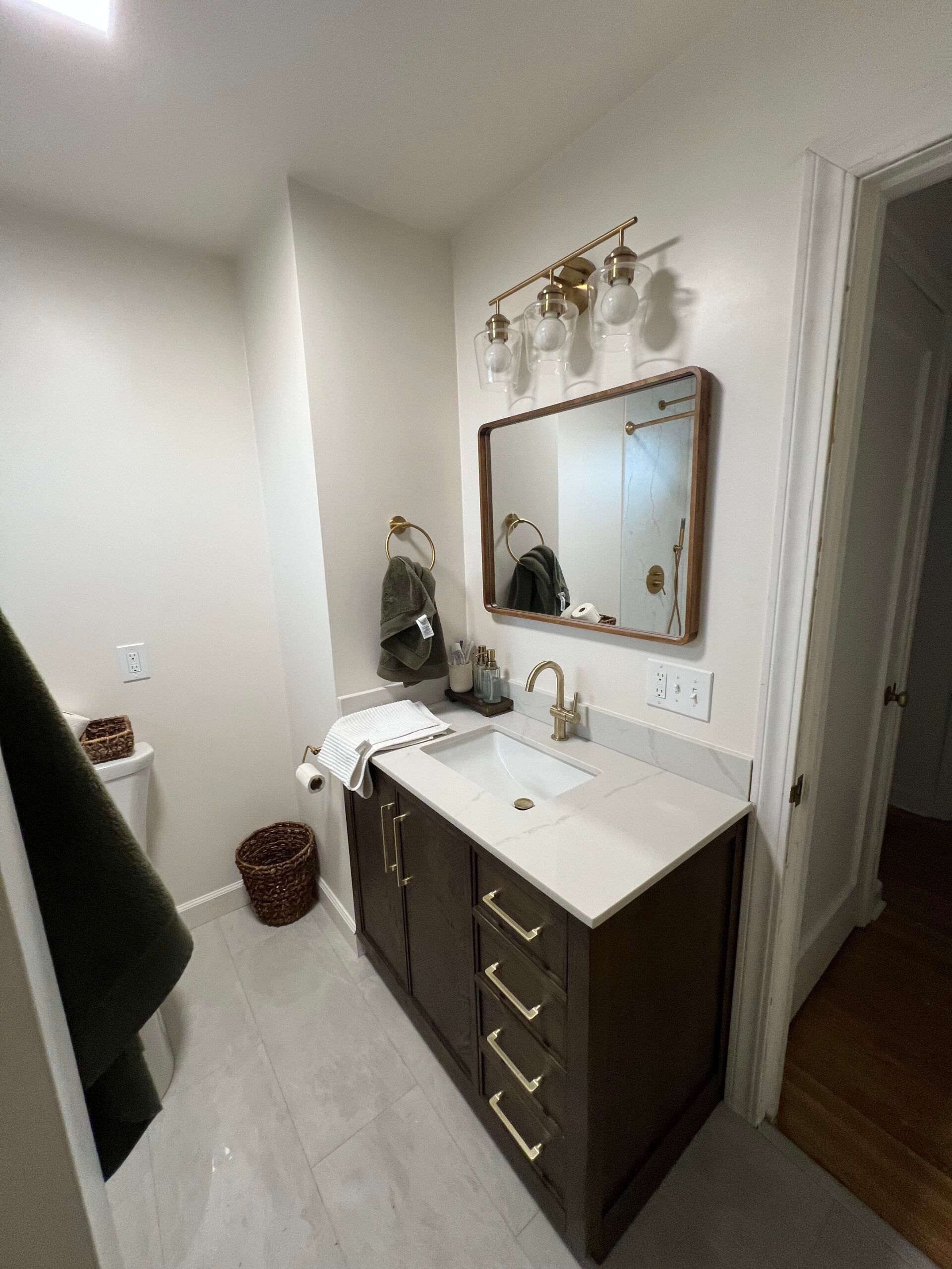 Compact bathroom vanity with dark wood cabinets, white countertop, mirror, and wall sconce in a narrow hallway area