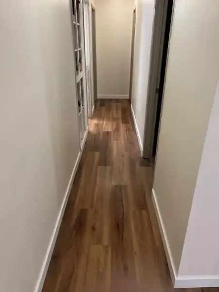 Narrow hallway with white walls and wood flooring, viewed toward an open doorway at the end.