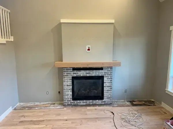 Empty living room with a central brick fireplace, beige walls, and unfinished wood floor with cables.