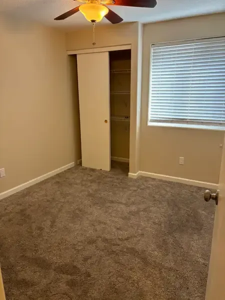 Empty beige bedroom with carpet, ceiling fan, open closet, and window blinds.