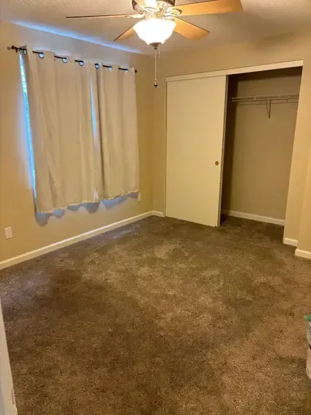 Empty beige bedroom with carpet, ceiling fan, window with curtains, and open closet doors.