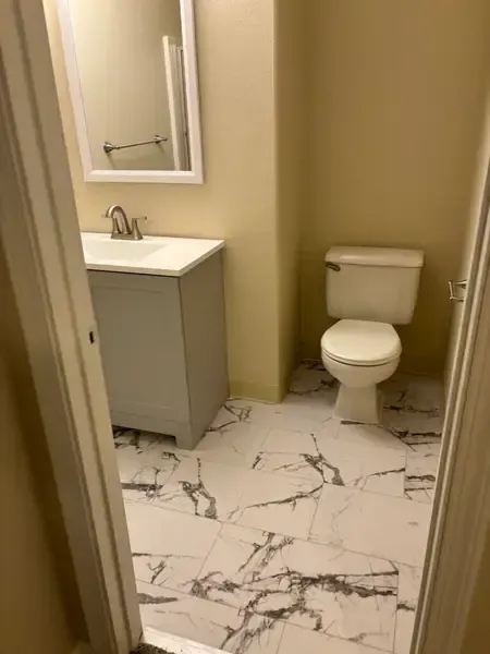Small bathroom with white vanity, mirror, toilet, and marble-pattern tile floor.