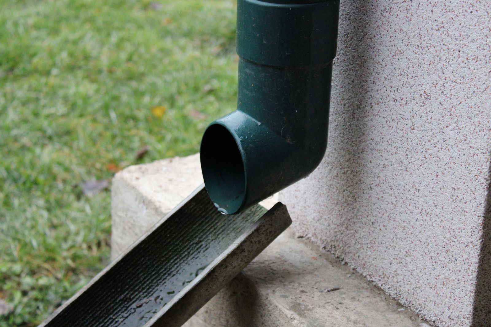 Green downspout draining rainwater into a black gutter beside a house foundation.