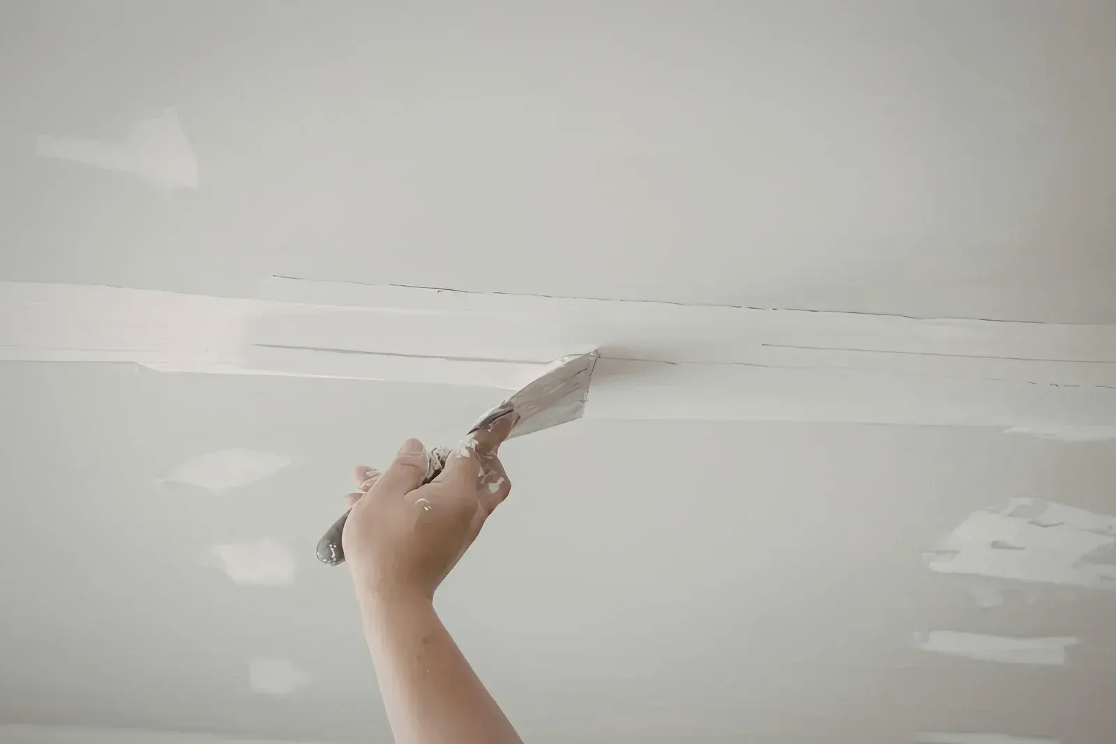Hand applying drywall compound to a white ceiling seam with a putty knife