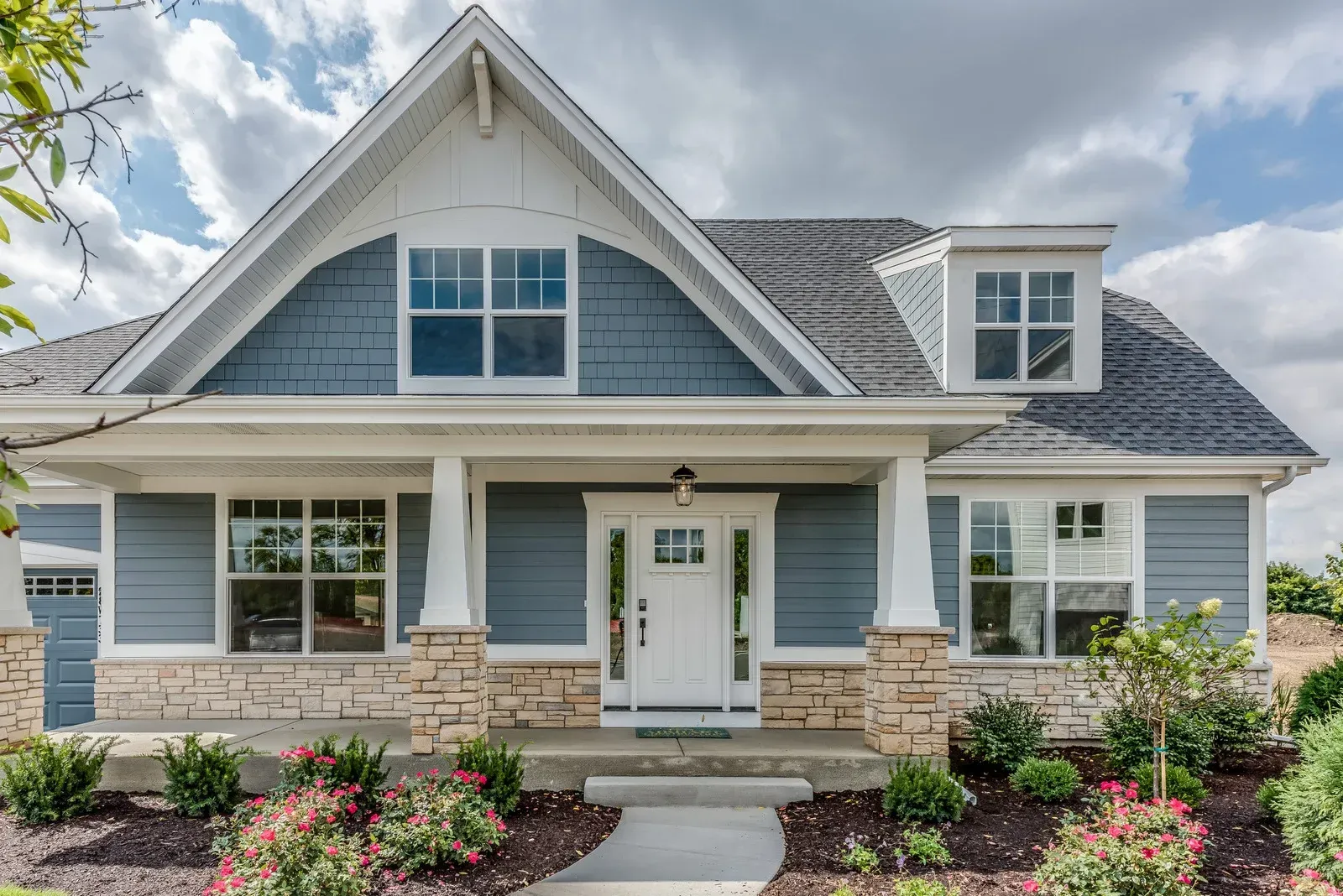 Blue-gray suburban house with white trim, front porch, stone columns, and landscaped flower beds