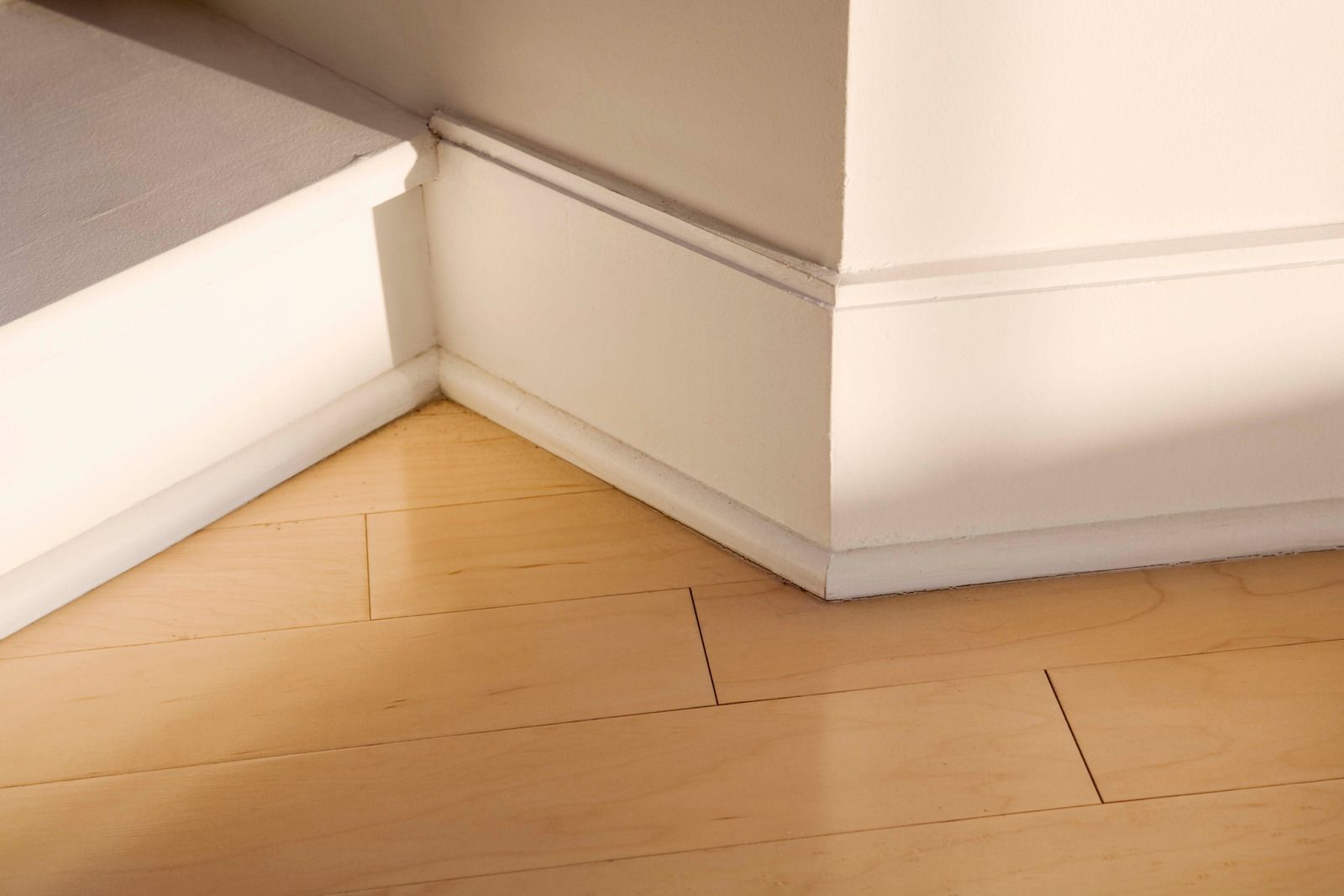 Wooden floor with a white baseboard and corner of a staircase, lit by warm sunlight.