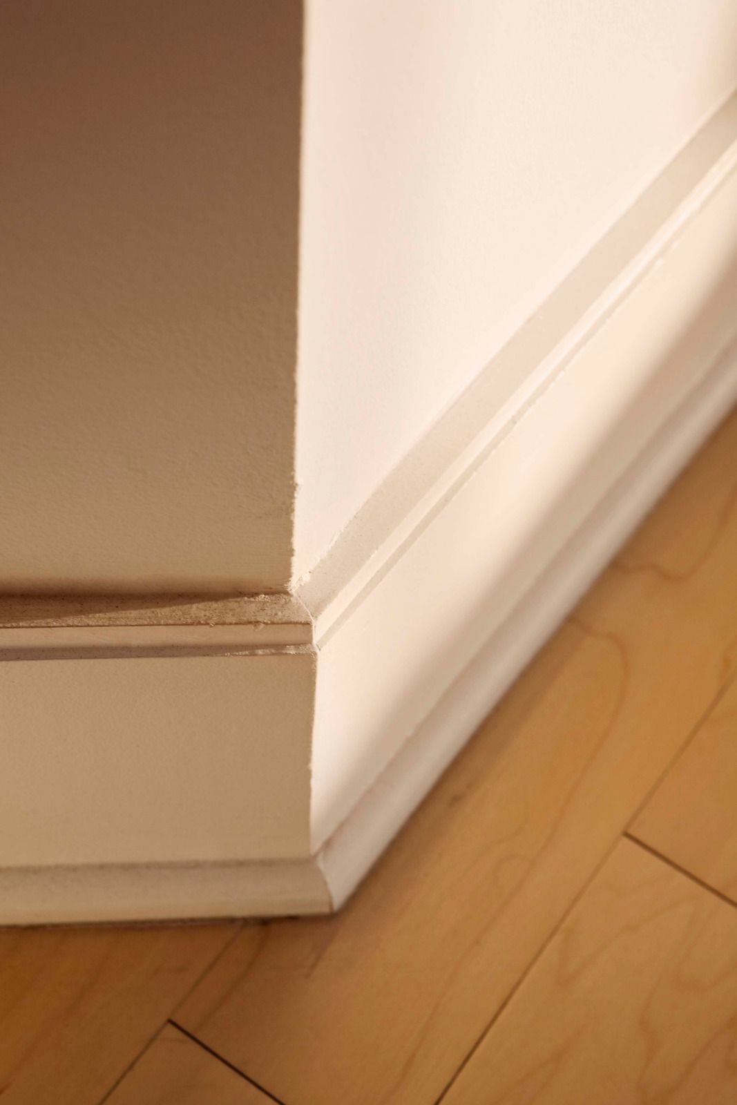 White baseboard trim on a beige wall above a light wood floor, showing a corner detail.