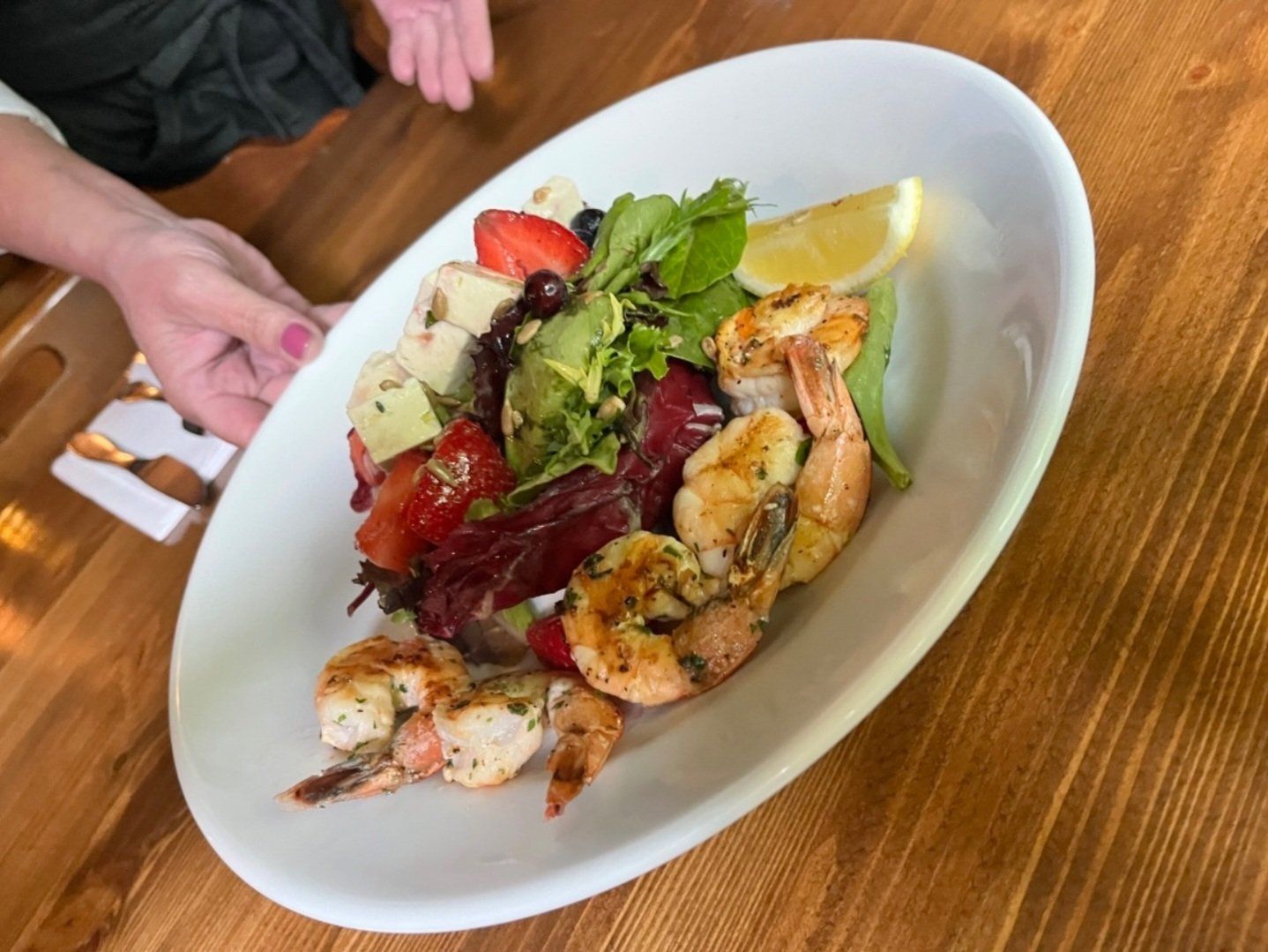 A person is holding a plate of shrimp and salad on a wooden table.