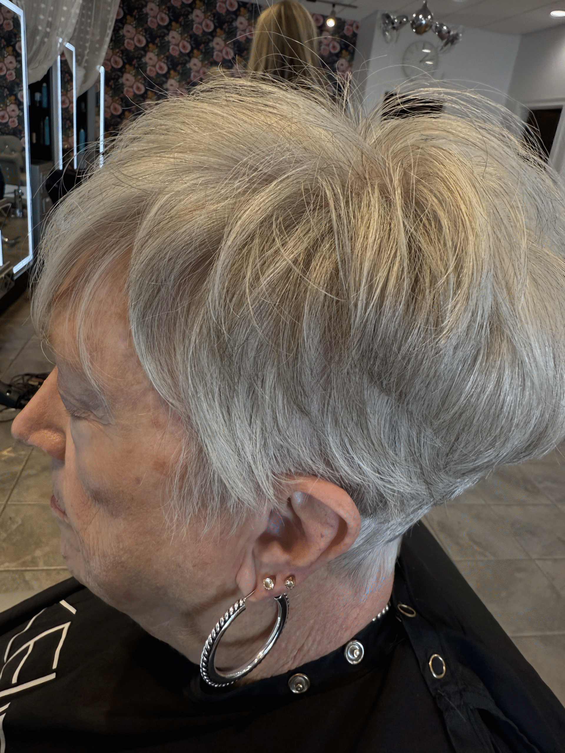 Side profile of a person with short silver hair in a salon chair, wearing hoop earrings and a black cape.