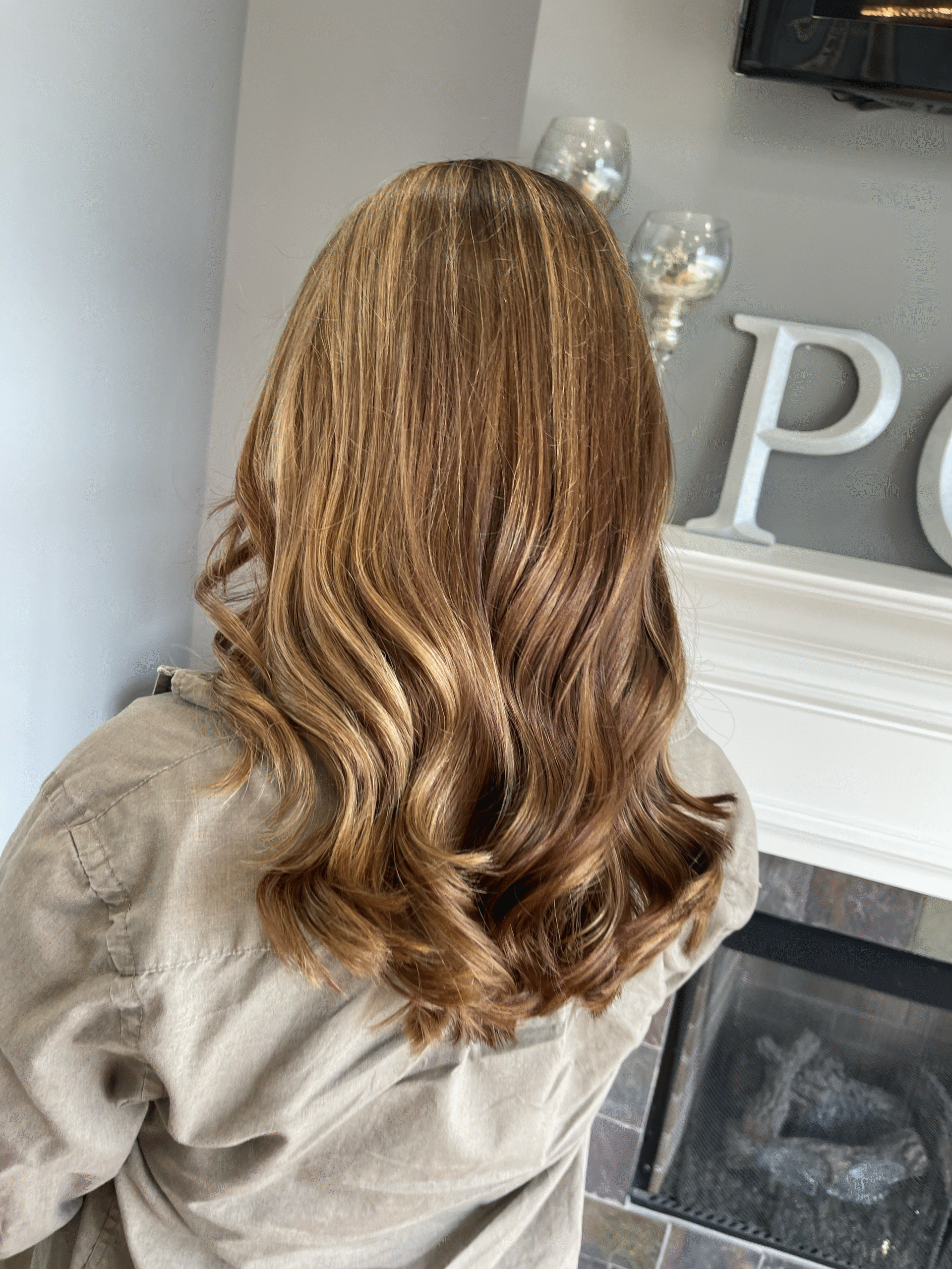 Woman with long, wavy light brown hair seen from behind in a living room near a fireplace.