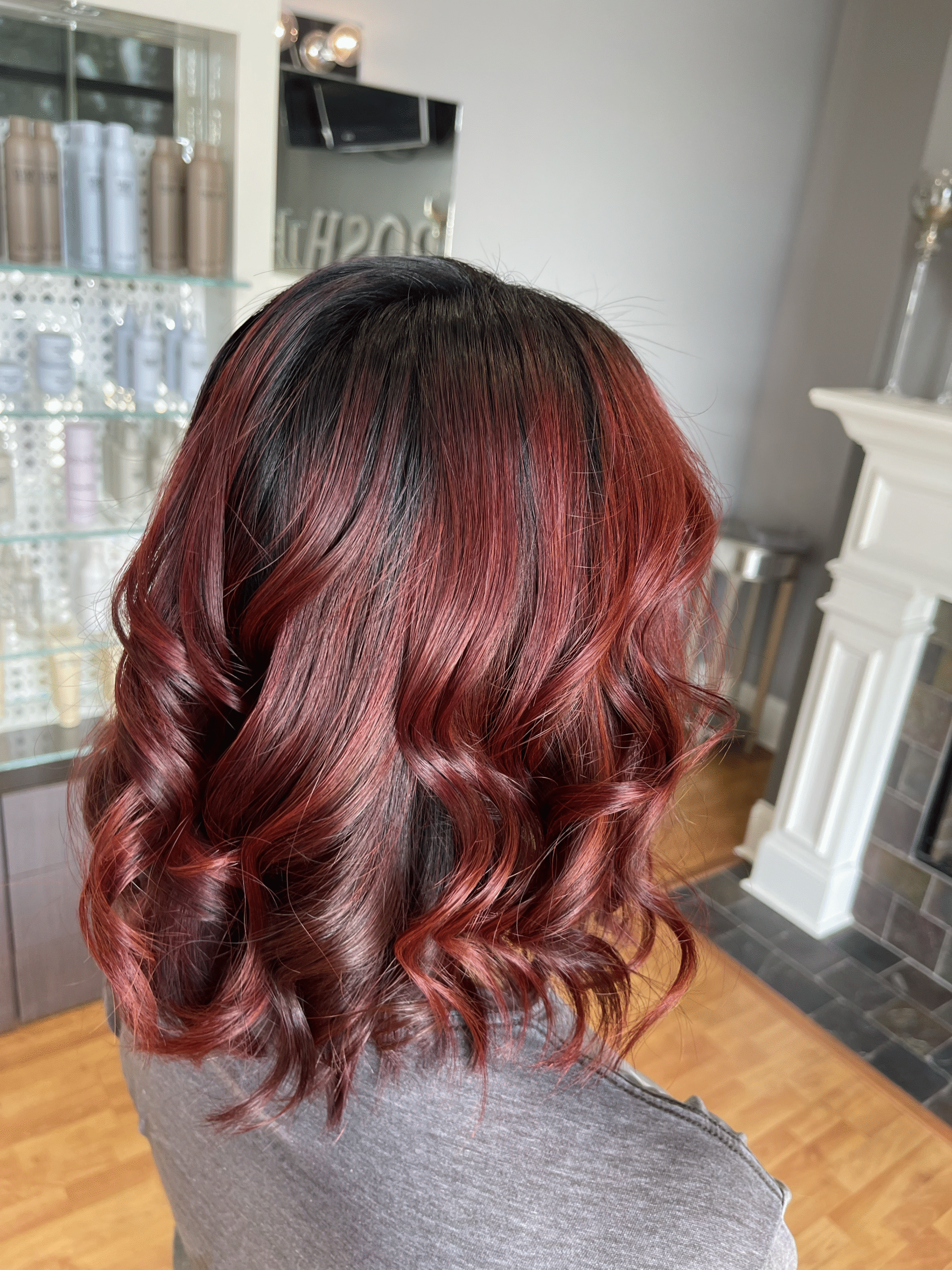 Shoulder-length burgundy curls with dark roots, viewed from behind in a bright indoor room.