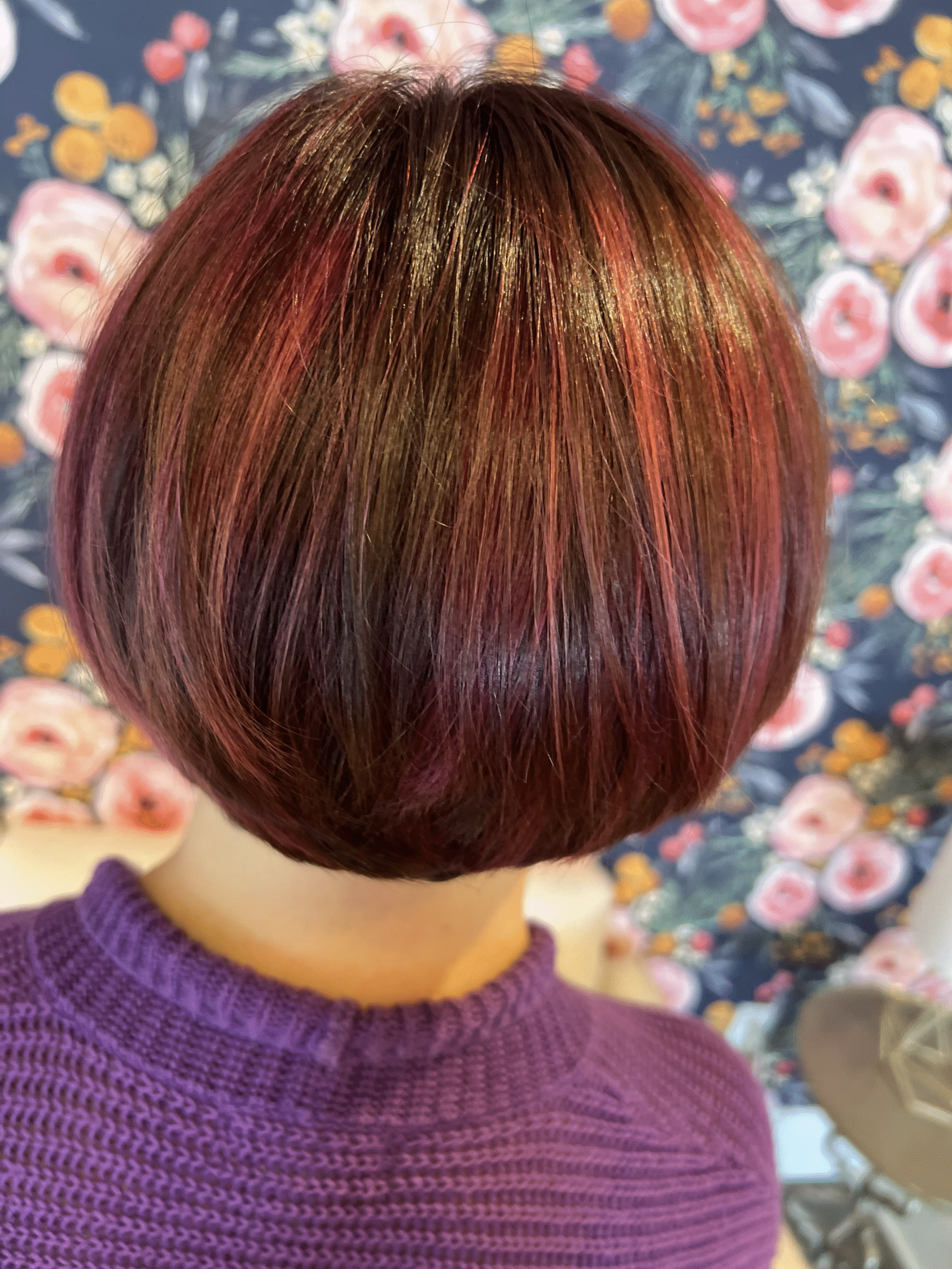Short burgundy bob haircut viewed from behind, with copper highlights against a floral wallpaper background