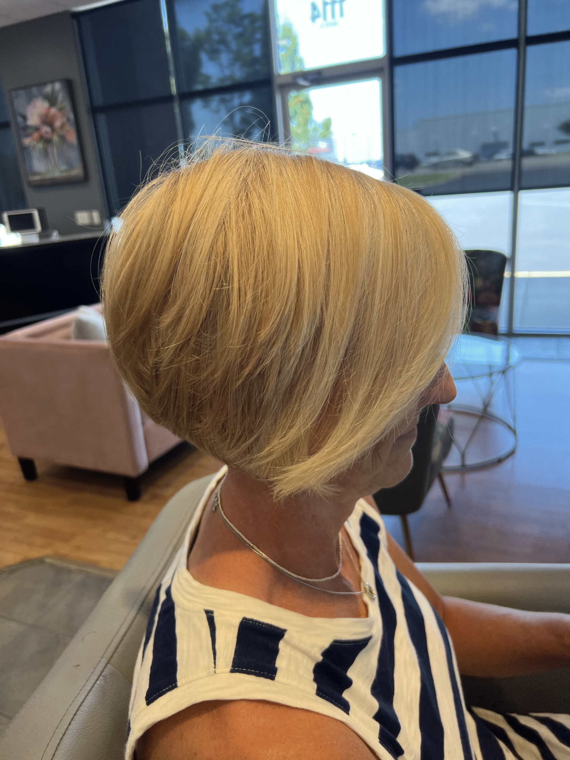 Person with short blonde hair seated in a salon, seen in profile near large windows.