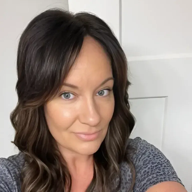 Portrait of a woman with dark wavy hair and a slight smile, wearing a gray patterned top indoors