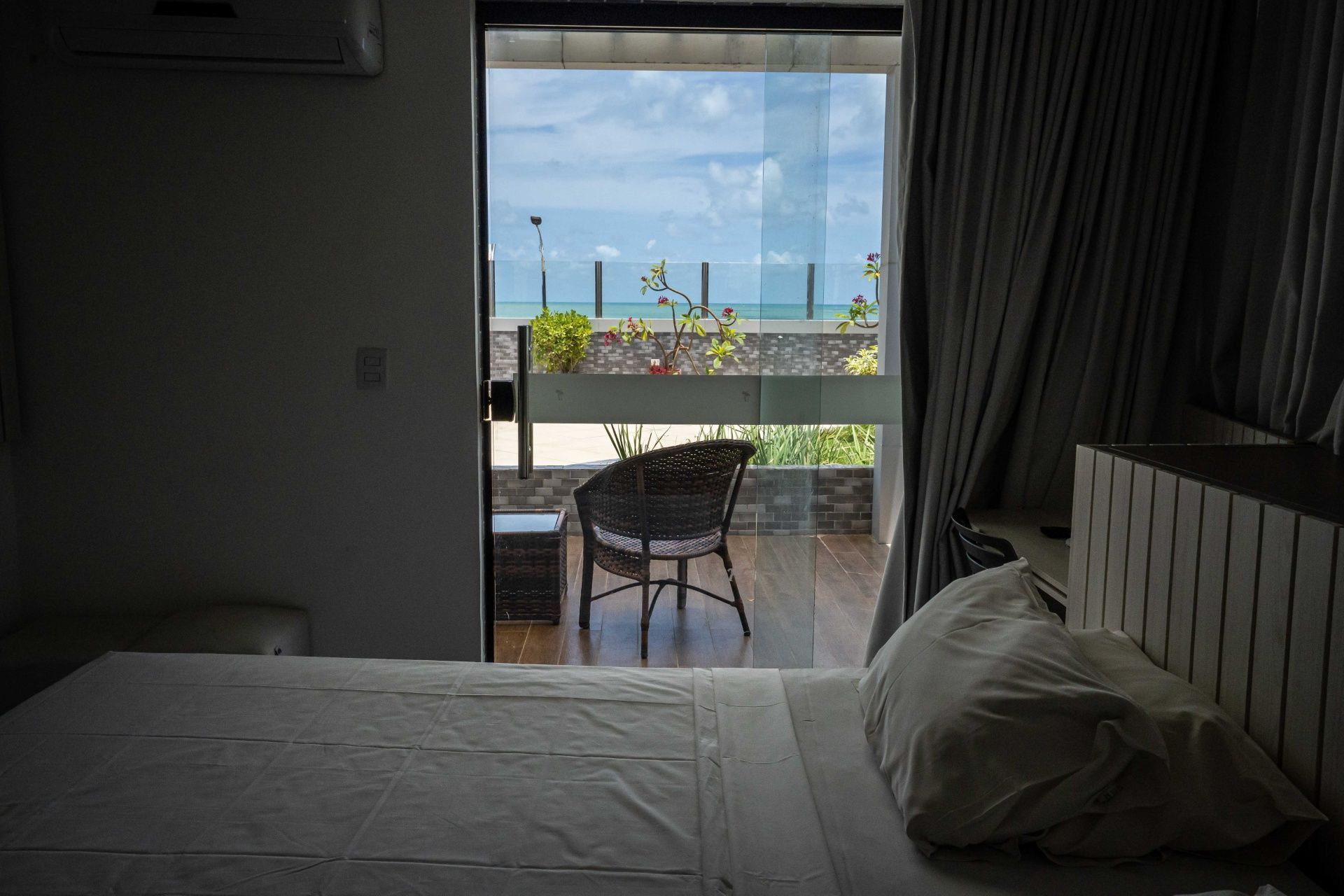 Quarto com cama e travesseiro, porta de varanda aberta e uma pequena cadeira com vista para o mar.