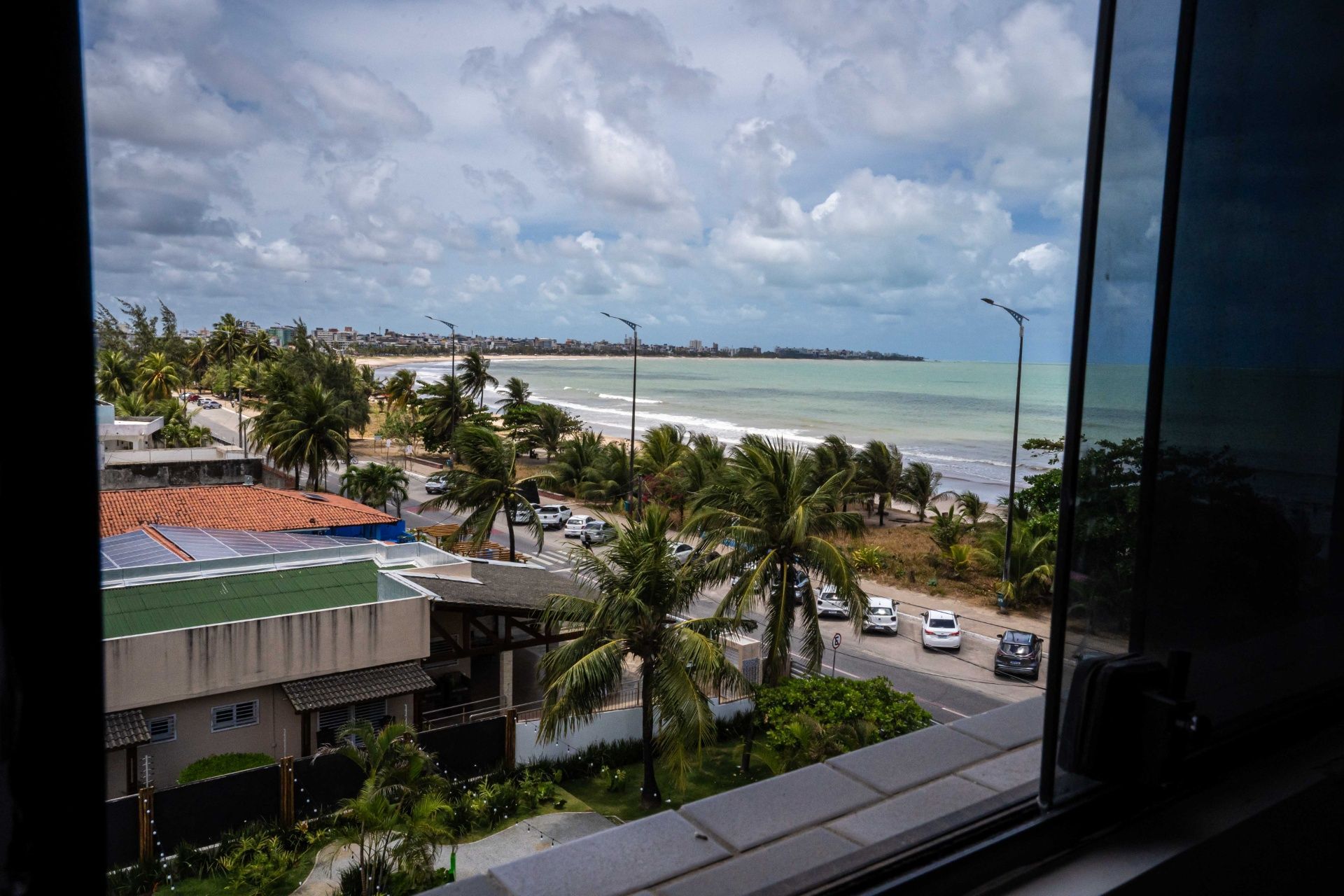 Vista da janela para uma costa tropical com palmeiras, prédios, carros e um céu azul nublado.