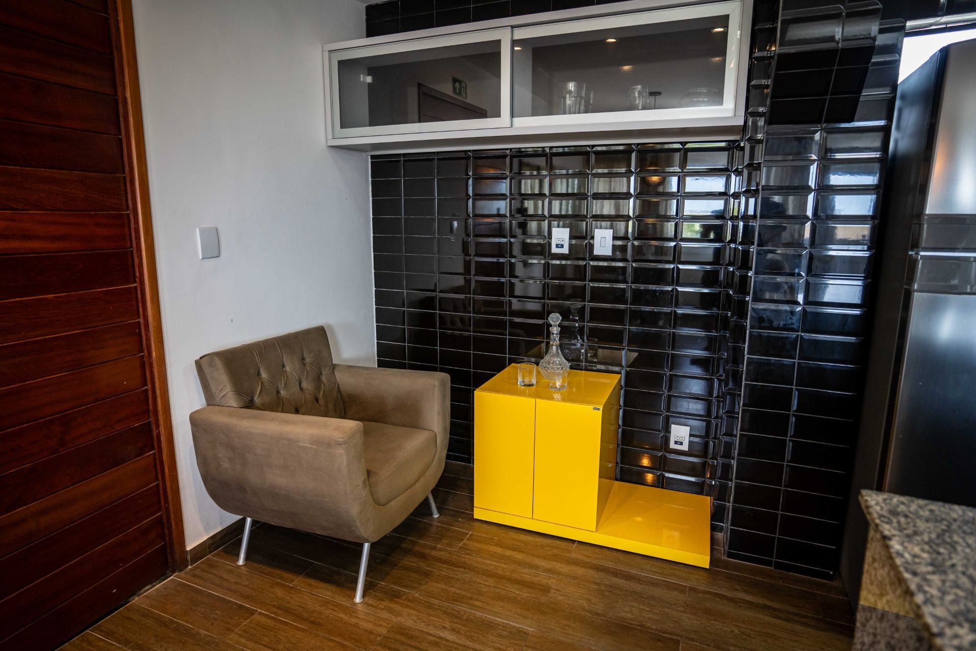 Sala de estar moderna de canto com cadeira bege, mesa lateral amarela e parede revestida com azulejos pretos.