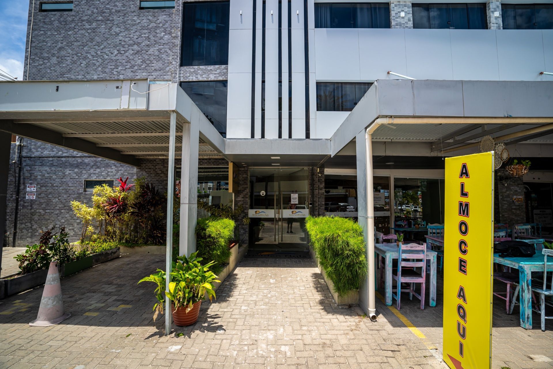 Entrada de um edifício moderno com vasos de plantas, caminho de azulejos e uma placa vertical amarela junto à porta.
