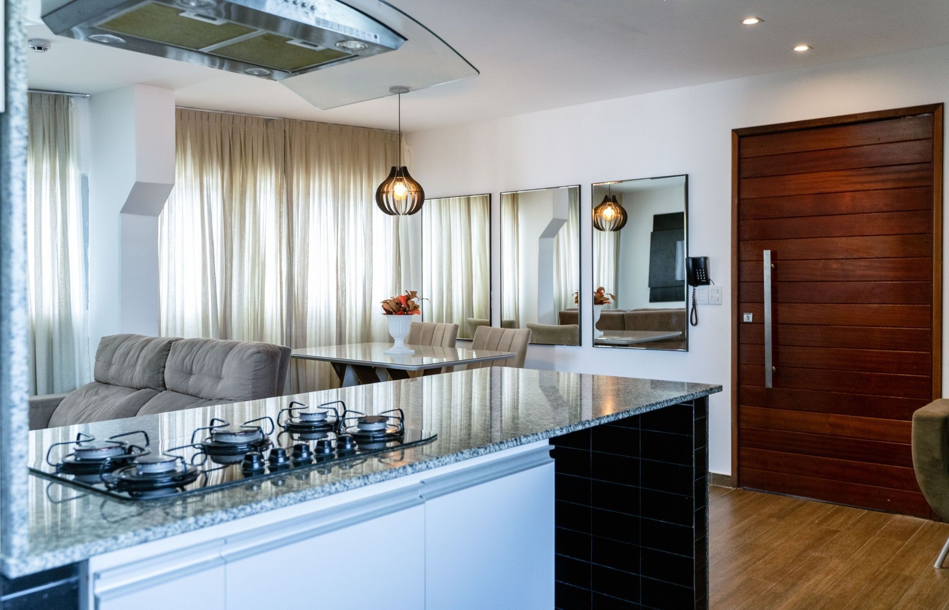 Cozinha moderna com ilha brilhante, sofá cinza, luminárias pendentes, espelhos e porta de madeira.