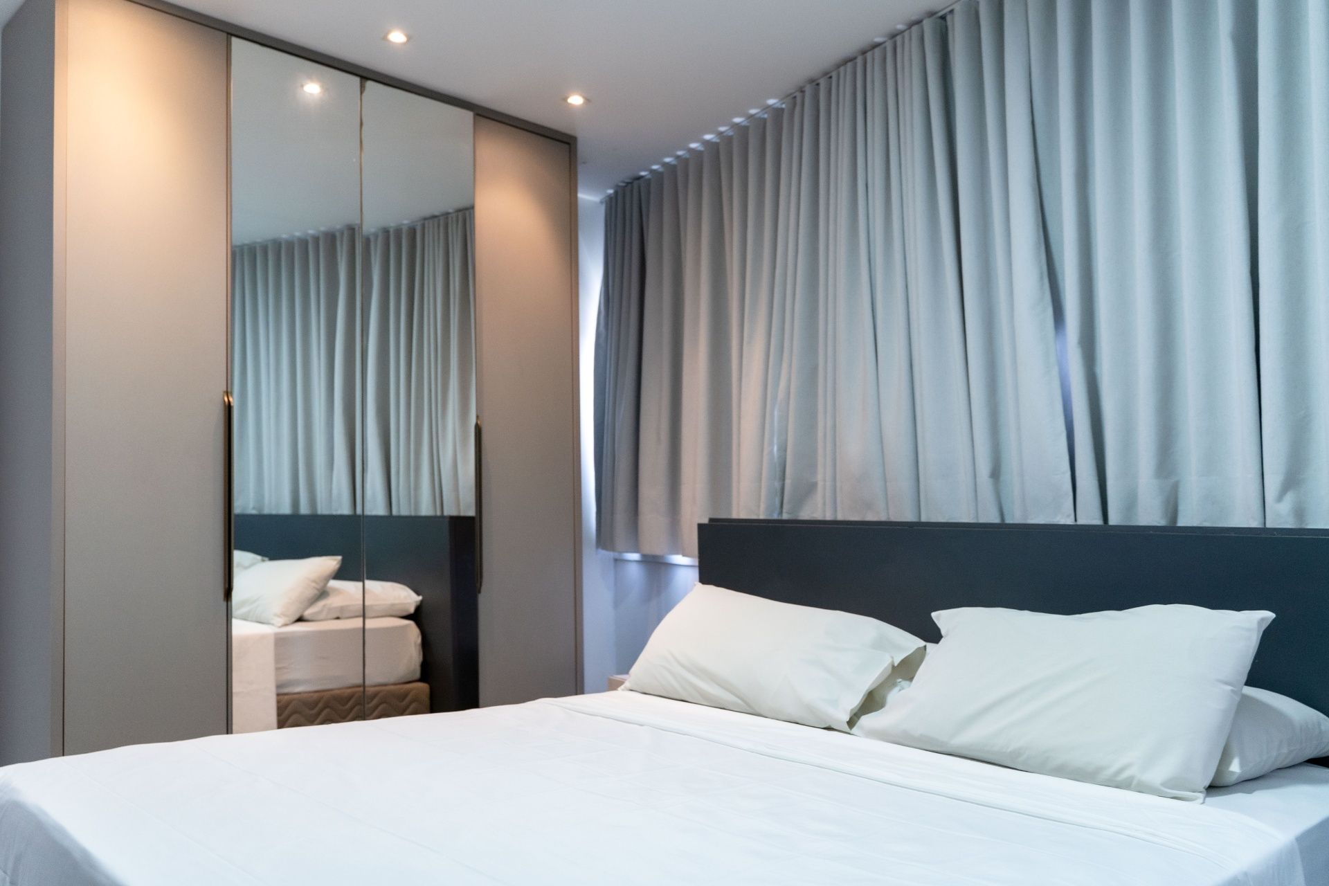 Quarto moderno com cama, guarda-roupa espelhado e cortinas cinza.