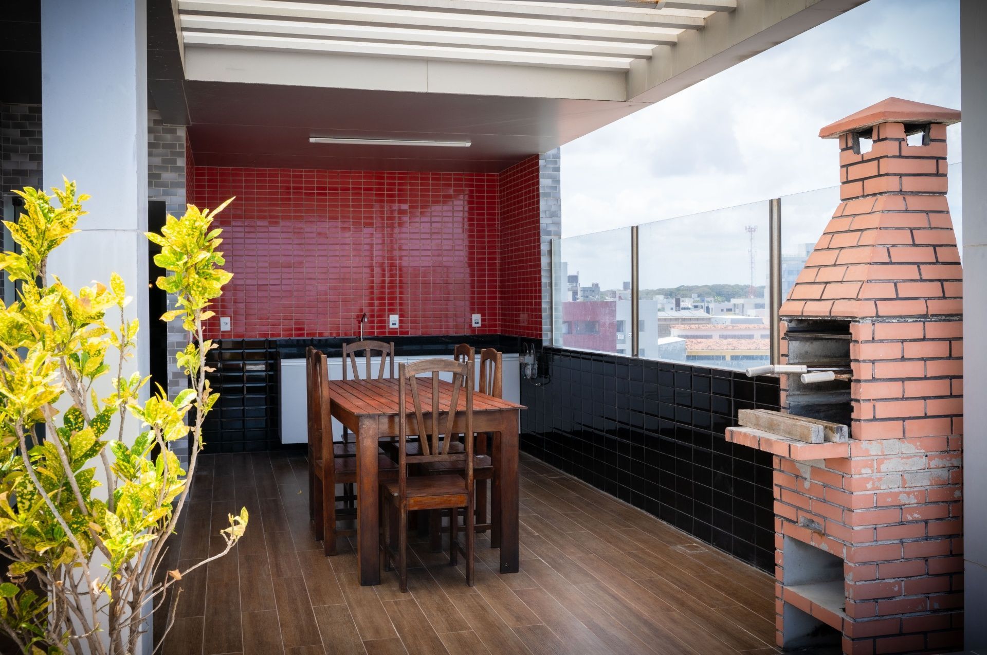 Área de jantar ao ar livre no terraço com mesa de madeira, parede vermelha, churrasqueira de tijolos e vista para a cidade.