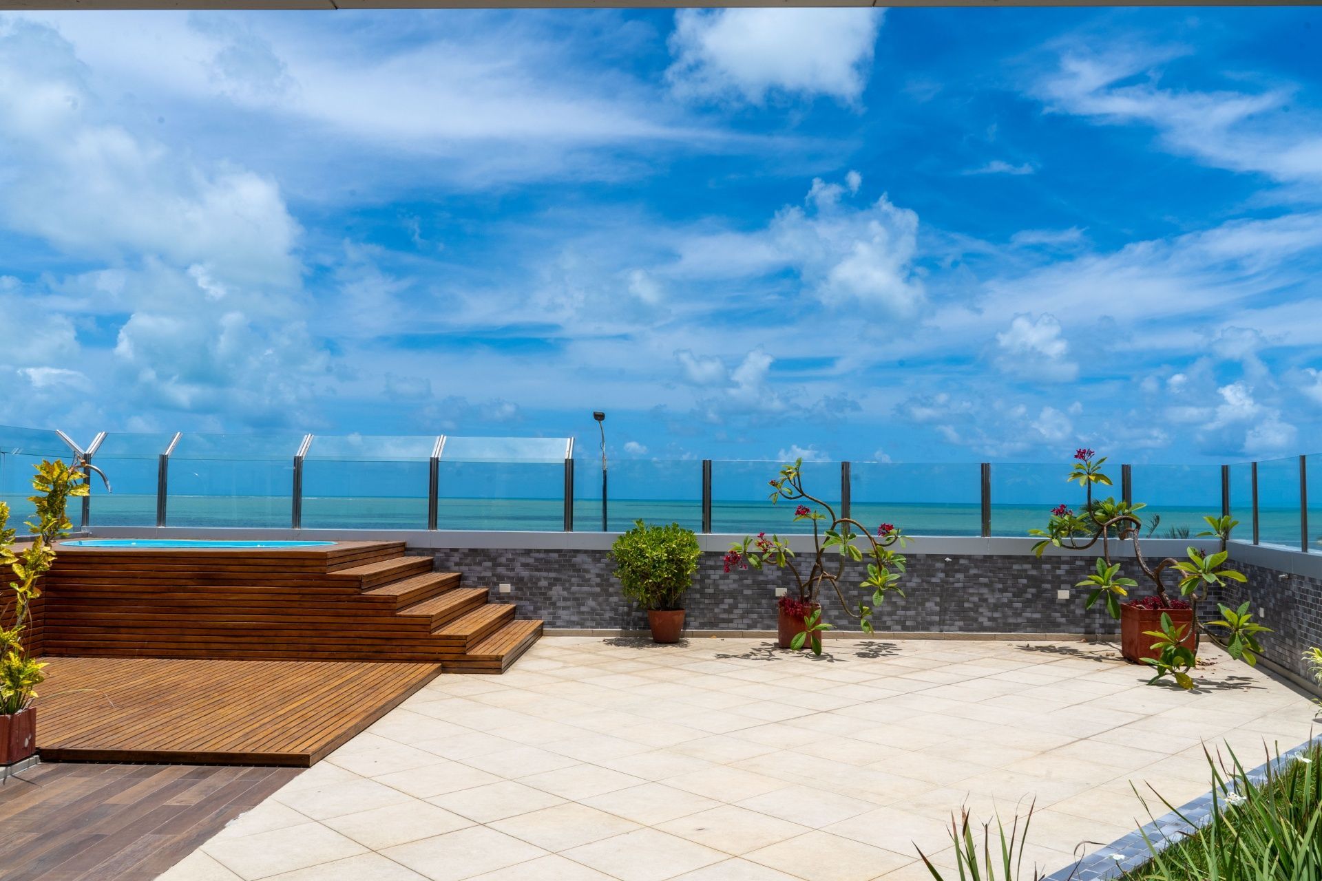Terraço à beira-mar com deck de madeira, plantas em vasos, turbinas eólicas e céu azul sobre o oceano.